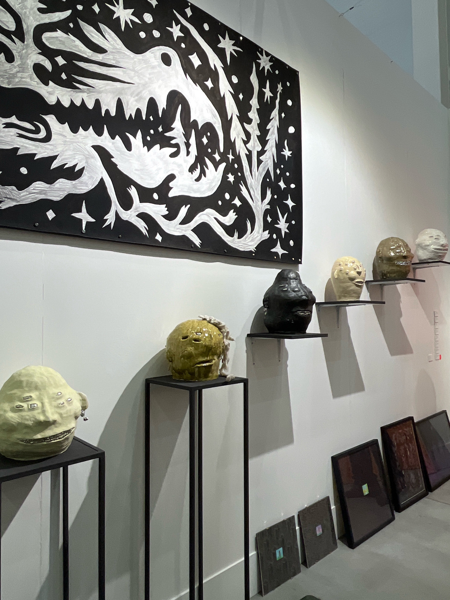 The image depicts an art exhibition in Khimki, Russia. On the wall, a large black and white mural of a stylized creature with sharp teeth and multiple eyes is displayed. Below the mural, on black shelves, are several ceramic sculptures of heads with exaggerated features, including multiple eyes and wide mouths. To the left of the shelves, two additional ceramic heads are displayed on black pedestals. In the foreground, leaning against the wall, are several framed artworks. The lighting suggests an indoor gallery setting.