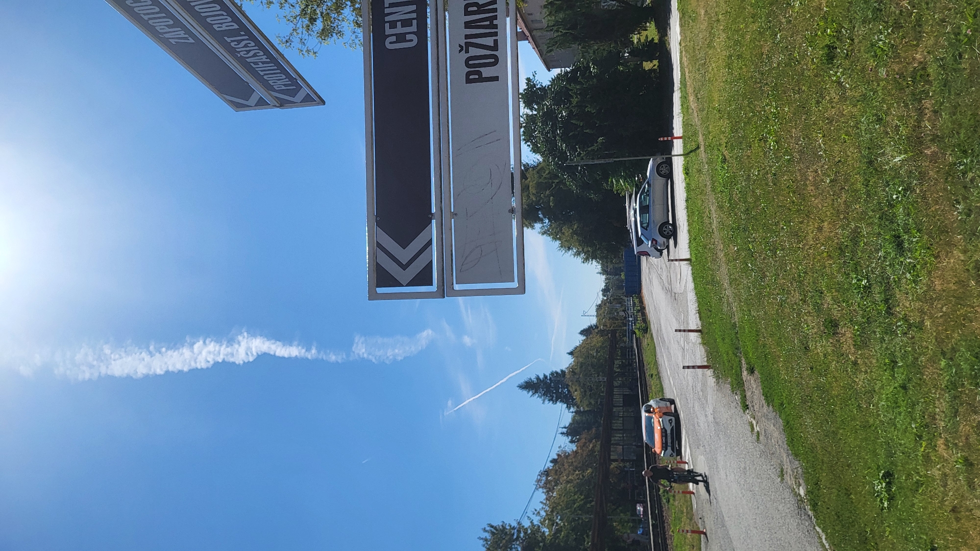 The image depicts a street scene in Prešov, Slovakia, on a sunny day. A tall, thin contrail stretches vertically across the bright blue sky, with another fainter contrail diagonally in the distance. A directional signpost with two arms is visible in the upper right corner, with the top arm pointing left and displaying text that appears to read "ZAPOTOC." The lower arm, also pointing left, has text that seems to say "PROTIFASIST. BOJOV." Below these, another signpost points left with a black background and white text reading "CENTR." Below that, a white sign reads "POŽIARI." In the foreground, a grassy area transitions to an asphalt road. On the road, a man on a bicycle rides past an orange and white car parked on the side. Further down the road, two silver cars are parked. Trees and buildings form the background. The scene appears to be near a railway line, indicated by tracks and a covered platform on the left side of the image.