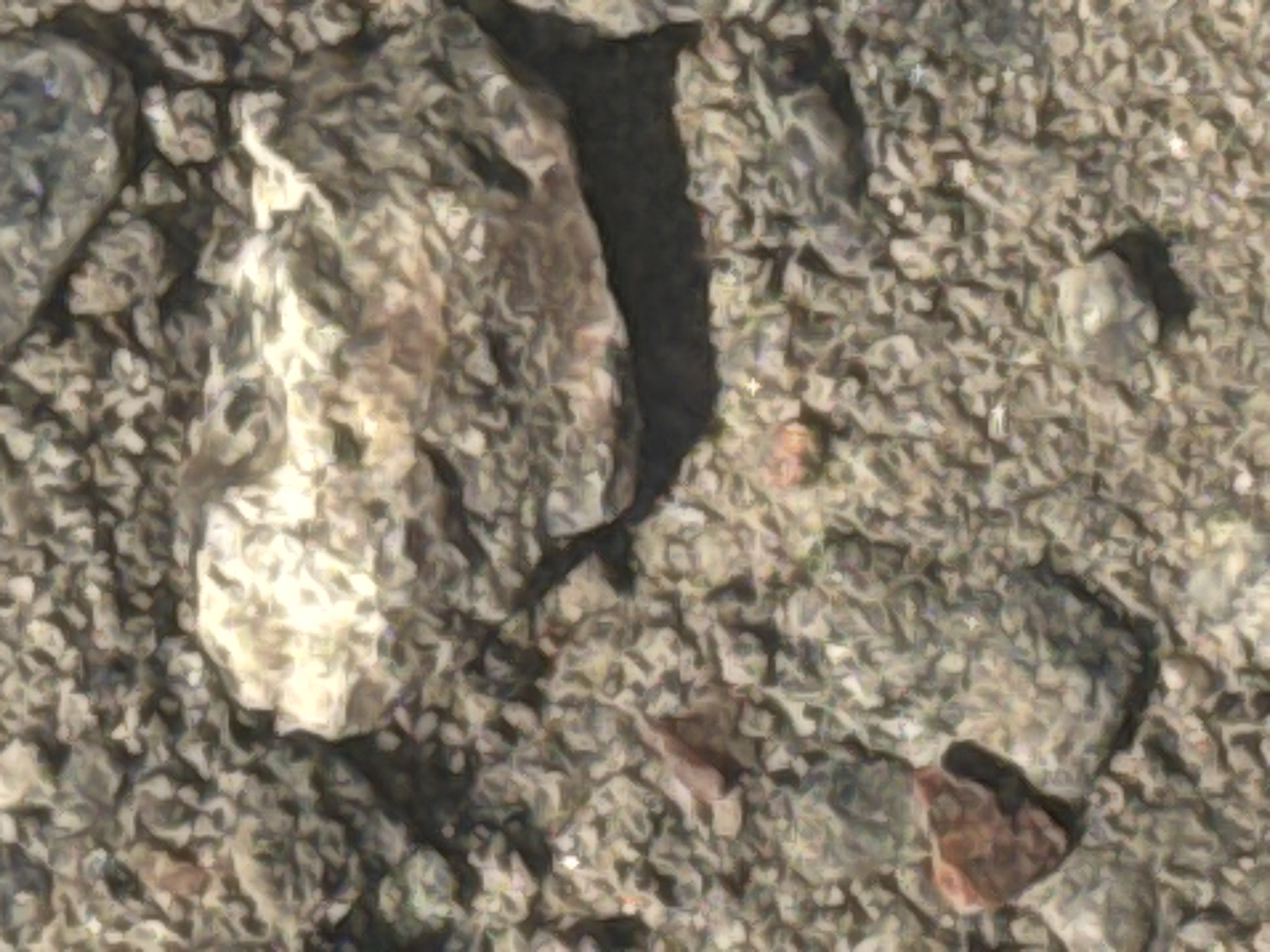 The image is a close-up, overhead view of a surface covered in various sized rocks and gravel. The dominant feature is a large, light-colored, somewhat flat rock positioned in the upper center of the frame. The surrounding ground is composed of smaller, darker grey and brown stones, interspersed with finer gravel. A few smaller, reddish-brown stones are visible, particularly in the lower left portion of the image. The lighting suggests an outdoor environment, possibly under direct sunlight, as evidenced by the highlights on the rocks and the shadows cast. The scene appears to be an unpaved area, such as a trail, a construction site, or a natural landscape. There are no people or discernible man-made structures in the image.