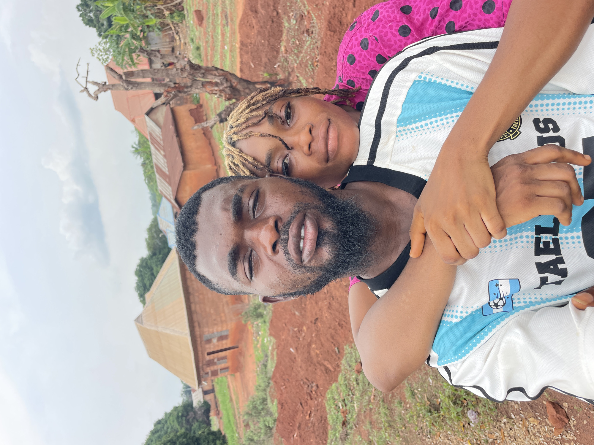 A photograph taken at Ogbe, Nigeria, shows a man and woman in an embrace. The man has dark skin, a full beard, and short dark hair. He is wearing a white and blue soccer jersey. The woman, slightly behind him, has light brown skin and dreadlocks with blonde highlights. She is wearing a pink top with black polka dots. Her arms are wrapped around the man. The background includes a dirt area, a few buildings with rusty roofs, and some greenery. The sky is overcast.