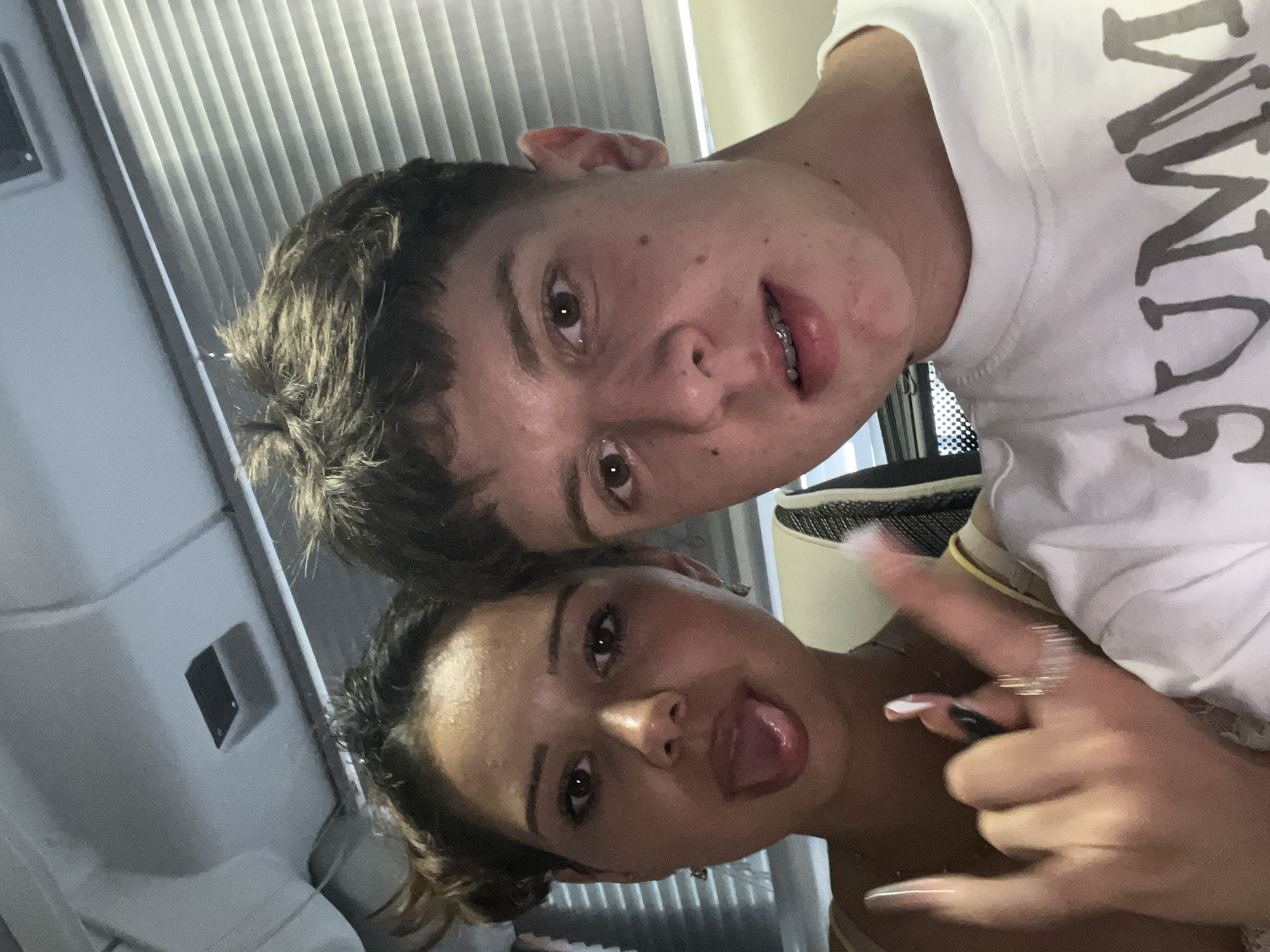 A selfie shows a young man and woman posing for the camera. The man is wearing a white t-shirt and has braces. The woman is sticking her tongue out and making a hand gesture. The background includes blinds and what appears to be the interior of a room. There is no information available to determine if this image was taken in Jesolo, Italy.