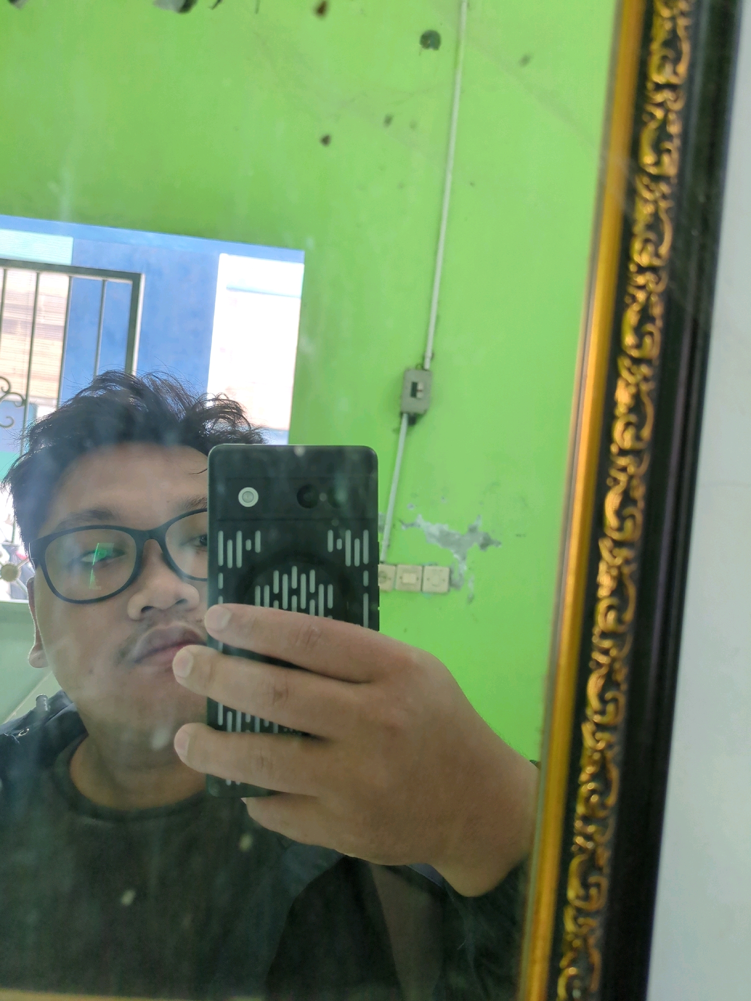 In a room with a bright green wall, a person is taking a selfie in a mirror. The person is wearing glasses and a dark t-shirt. They are holding a black smartphone in their right hand, with the phone's screen facing the mirror. The background in the mirror reflection shows a blue doorway with bars and white curtains. A decorative gold and black framed mirror is visible on the right side of the image. The scene appears to be indoors, possibly in a residential setting. The image was taken in Gumpang, Indonesia.