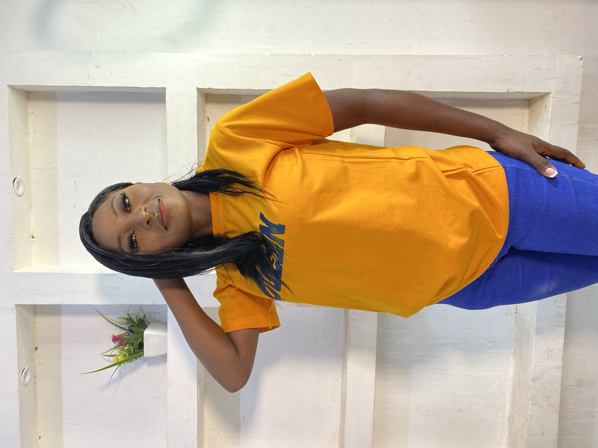 A Black woman is posing for a photo in front of a white wall with recessed shelves. She is wearing a bright orange t-shirt with blue lettering that appears to read "CLEAN" and blue jeans. Her right hand is behind her head, and she is looking towards the camera with a slight smile. One of the shelves on the wall contains a small white pot with artificial green, yellow, and pink flowers. The scene appears to be indoors. There are no visual cues to specifically place this image in Jos, Nigeria.