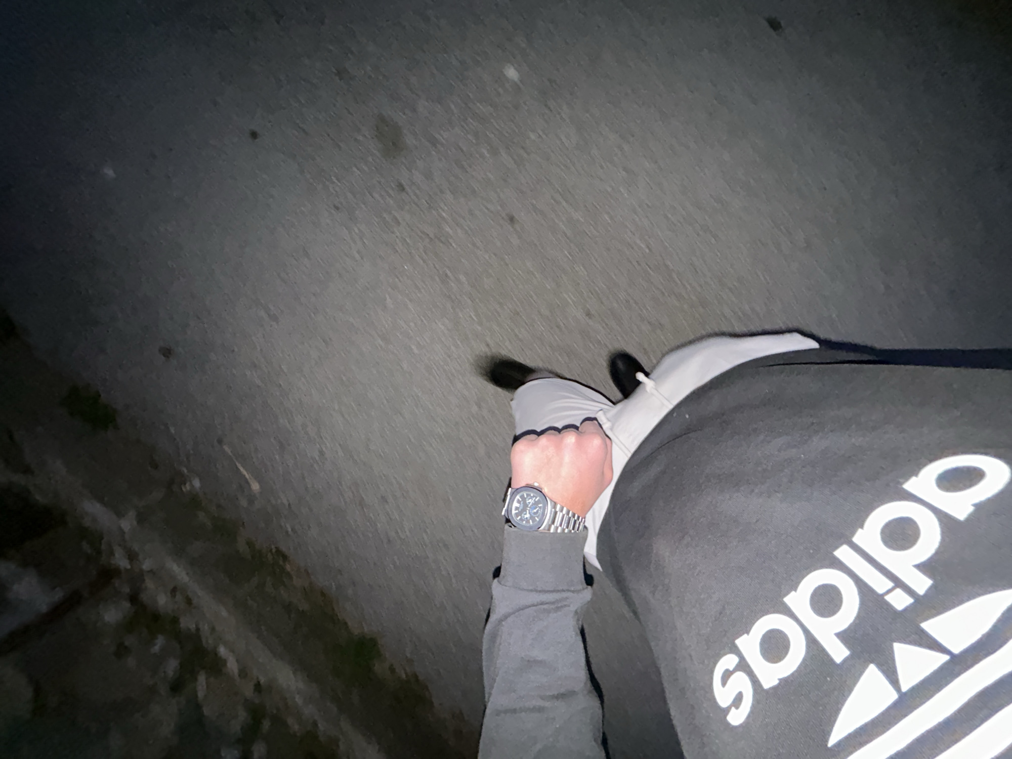 A first-person view shows a person standing on a paved surface, possibly a sidewalk or road, at night. The person is wearing a gray Adidas sweatshirt and light gray pants. A wristwatch with a metal band is visible on the person's left wrist. The ground surface is uneven, with some grass and vegetation visible on the left side. The location cannot be independently verified as Zlatograd, Bulgaria, from the visual evidence.
