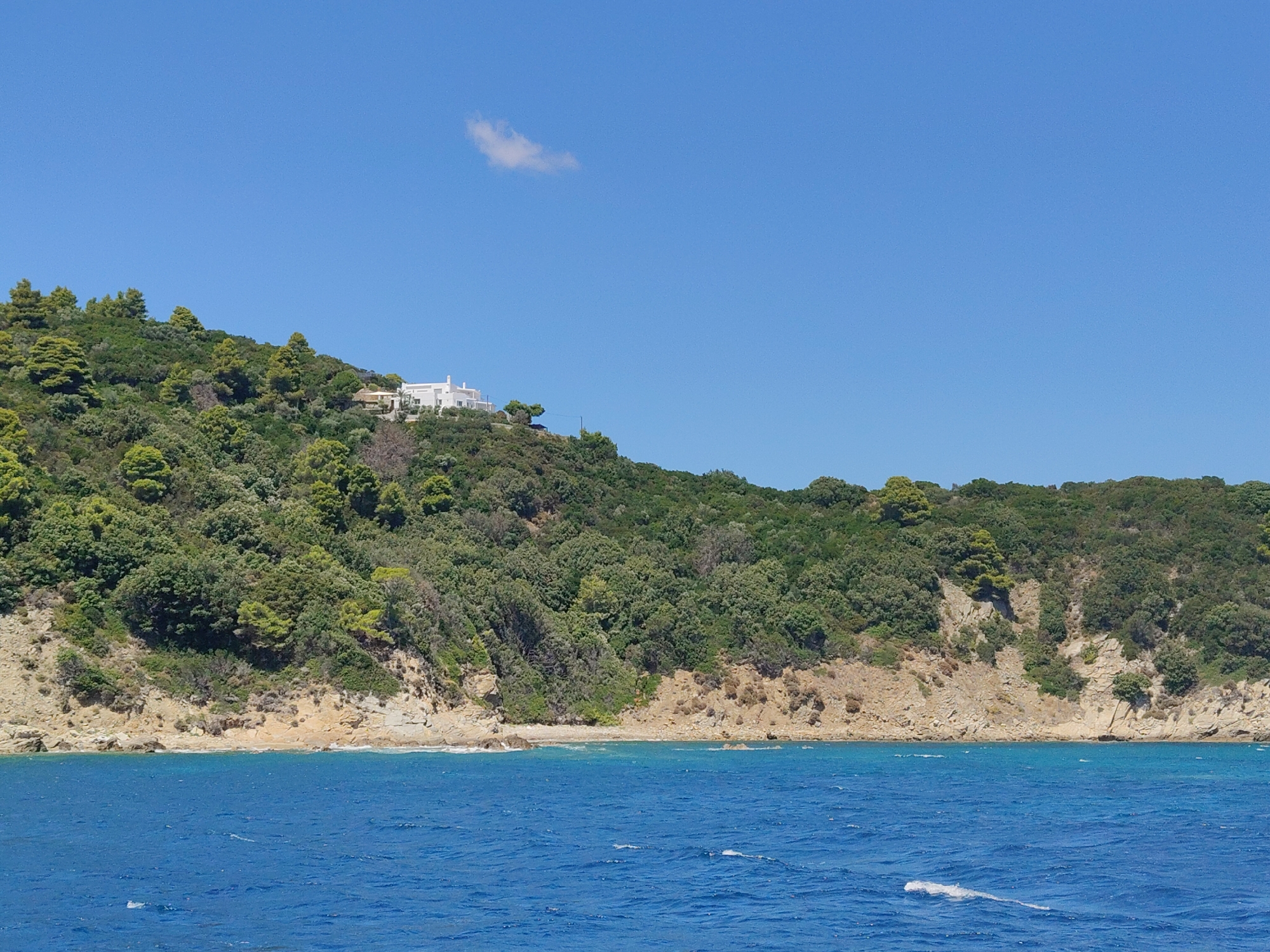 A serene coastal landscape unfolds under a clear blue sky, featuring a verdant, tree-covered hillside that slopes down to a rocky shoreline meeting the vibrant blue sea. Perched atop the hill is a white, modern house with multiple sections and a flat roof, overlooking the water. A single, wispy cloud floats in the upper portion of the sky. The image captures a tranquil day in Achladias, Greece, with the natural beauty of the island amplified by the presence of the elegant villa.