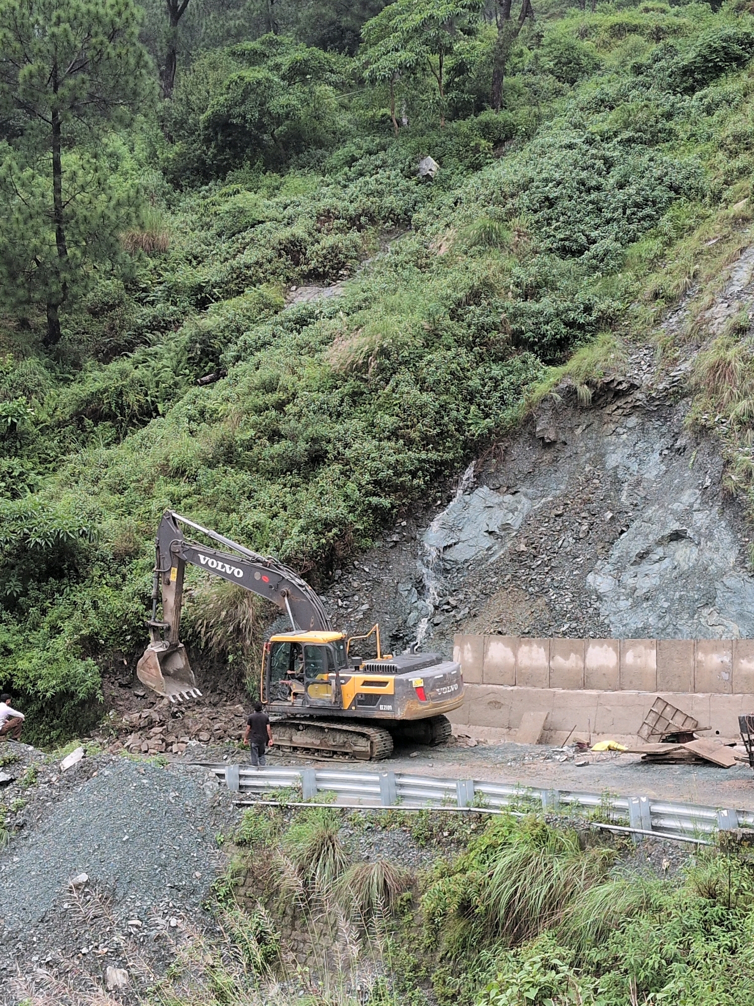 In Gumma, India, an excavator is seen working on a steep, tree-covered mountainside. The excavator, a Volvo model with the number "EX2100" visible on its side, is positioned on a dirt path alongside a protective barrier and concrete blocks. The machine's bucket is lowered, appearing to scoop or move debris from the slope. To the left of the excavator, a person wearing a dark shirt stands facing away from the camera. Further left, another individual, dressed in lighter clothing, is crouched down. The environment is characterized by lush green vegetation and rocky terrain, suggesting a construction or maintenance project on a mountain road.