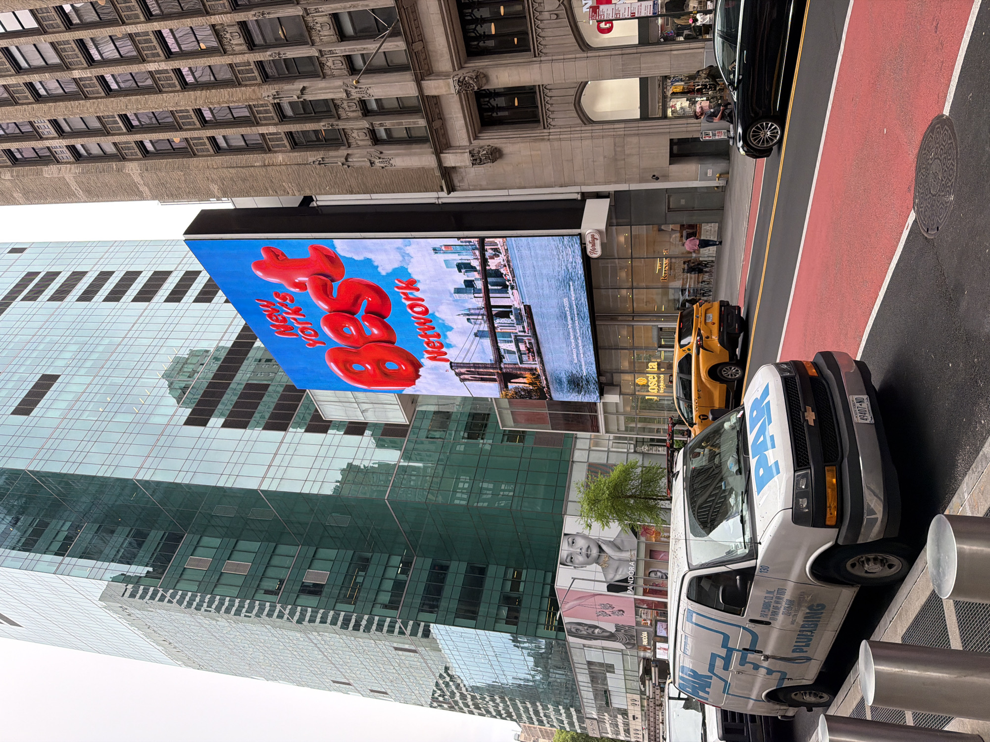 Here is a description of the image:
The image is an outdoor shot taken in New York City, USA. It features a prominent digital billboard advertising "New York's Best Network" with red lettering and imagery of the city skyline. The billboard is attached to a modern building with a glass facade reflecting the surrounding structures.
A white van with blue "PAR Plumbing" lettering is parked on the street. A yellow taxi cab is visible behind the van. Pedestrians can be seen walking on the sidewalk in front of stores. The street is marked with red lanes and a sewer cover is visible on the edge of the roadway. Older buildings with intricate architectural details are present in the background, contrasting with the more modern architecture.
