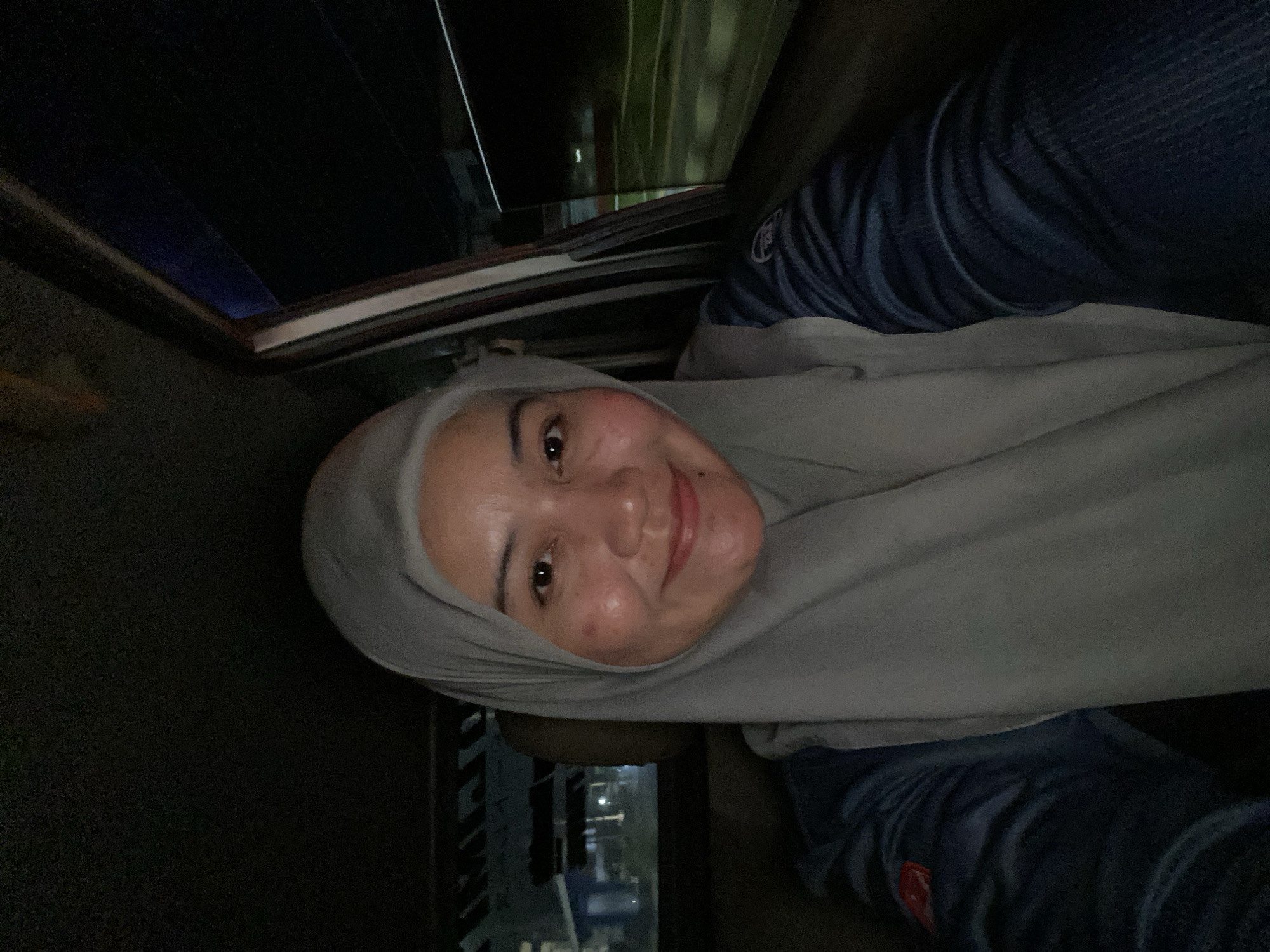 The media file shows a close-up selfie of a woman wearing a light gray hijab and a dark blue shirt. She is looking at the camera with a slight smile, her face illuminated by soft lighting. The background is dark, suggesting an indoor setting at night, possibly in a car due to the visible car interior elements like the window frame and what appears to be a sunroof. Her shirt has a visible logo on the sleeve, which is red and white. The image is oriented sideways. The location context provided is Jawa, Indonesia. There is no visible text in the image itself.