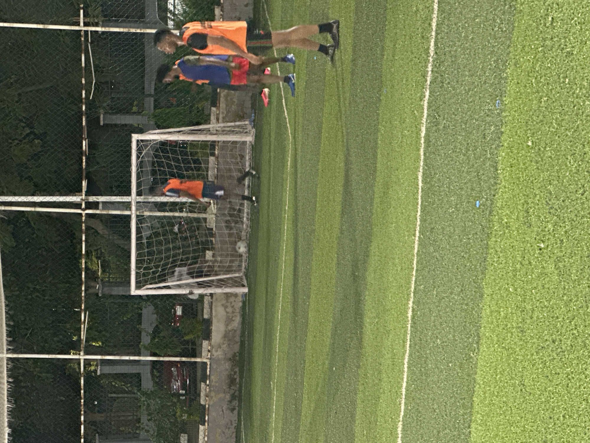 In Abuja, Nigeria, at an outdoor artificial turf soccer field, three men are practicing. The field is illuminated by overhead lights, casting shadows and highlighting the green turf marked with white lines. A soccer goal with a net stands in the background, positioned behind a chain-link fence and some vegetation. One man, wearing an orange vest and dark shorts, is near the goal. Two other men, one in a blue vest and red shorts and the other in an orange vest and black shorts, stand further to the right on the field. The man in the blue vest is facing away from the camera, while the man in the orange vest is facing the camera and looking to the side. The scene appears to be captured at dusk or night, with artificial lighting contributing to the overall illumination.
