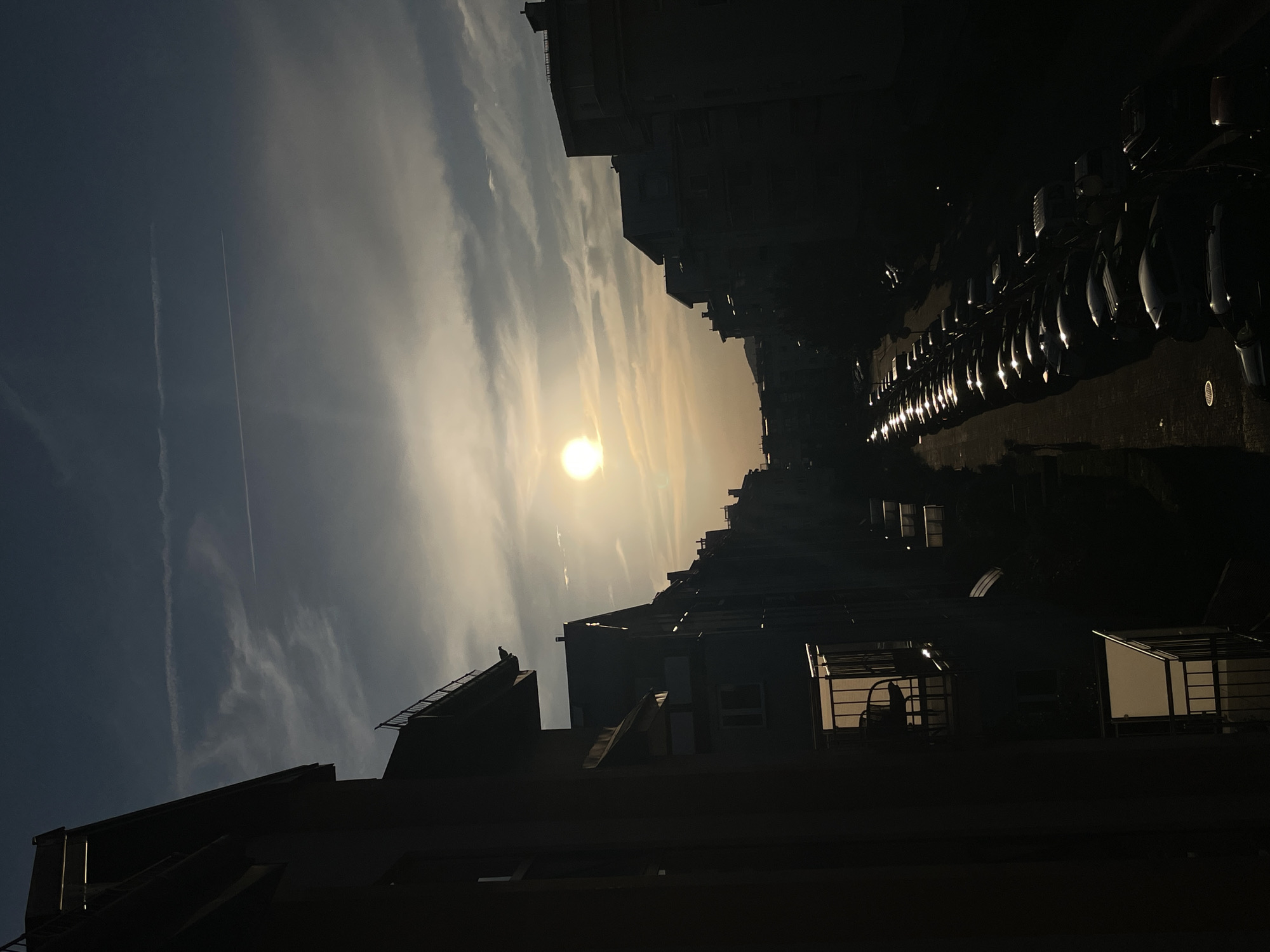 The image depicts a city street scene at sunset, with buildings silhouetted against a hazy sky. The sun, low on the horizon, casts a warm glow, illuminating wispy clouds and creating a dramatic, low-light effect. The foreground is dominated by the dark shapes of buildings, their architectural details obscured by shadow. In the middle ground, a row of parked cars is visible, their surfaces reflecting the ambient light. The overall impression is one of urban tranquility and the ephemeral beauty of the setting sun.
The orientation of the image is rotated 90 degrees clockwise. The time of day is clearly sunset, suggested by the low angle of the sun and the warm, diffused light. The sky appears somewhat hazy, possibly due to pollution or atmospheric conditions. The presence of buildings and parked cars indicates an urban or suburban environment. No people are visible in the image, and the atmosphere is one of quiet observation. The image is characterized by strong contrasts between light and shadow, with the bright sun and illuminated clouds juxtaposed against the dark silhouettes of the urban landscape. The scene evokes a sense of calm and introspection as the day draws to a close.