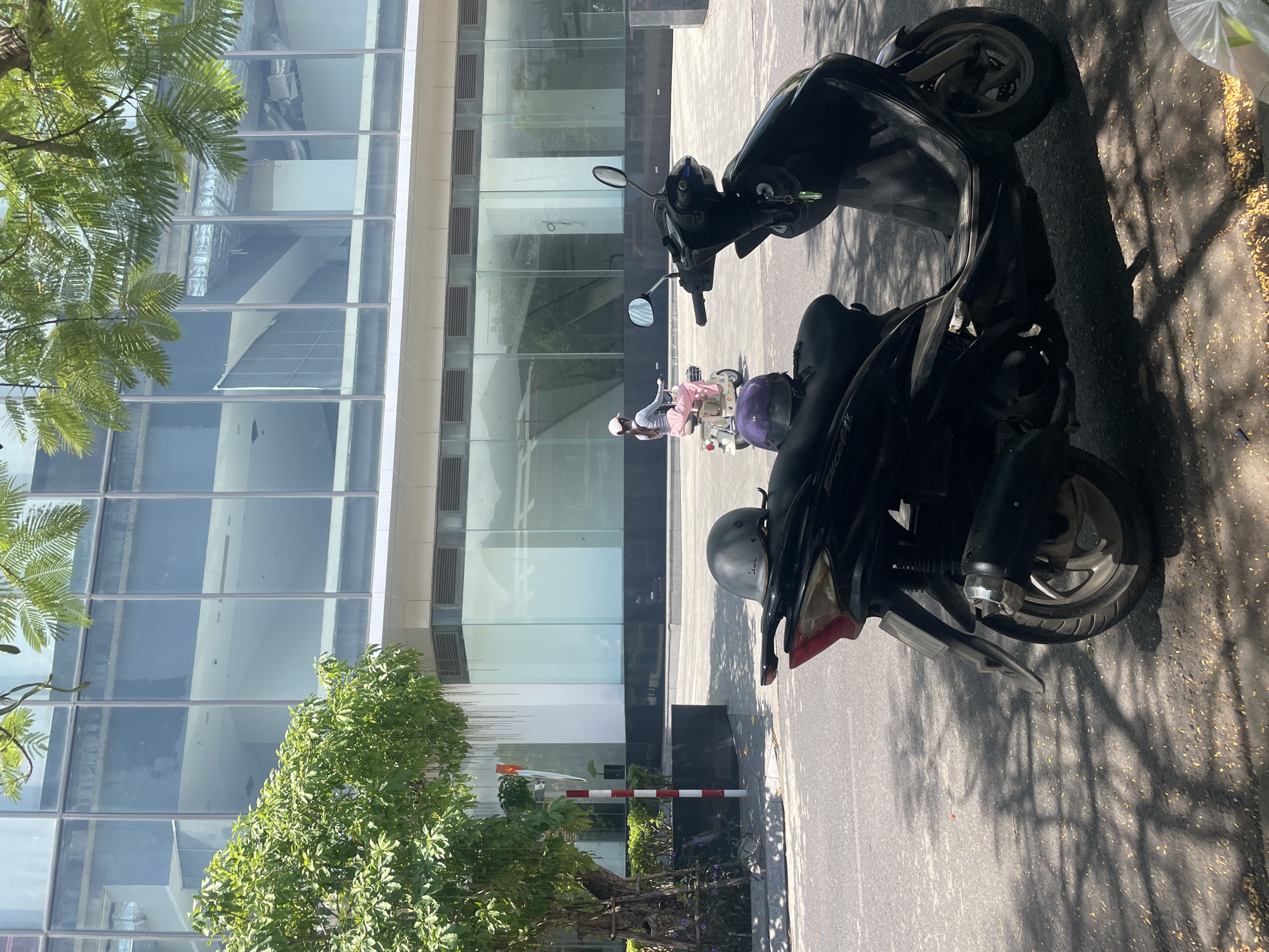 The image presents a sunlit urban scene in Bac Nha Trang Ward, Vietnam, characterized by modern architecture and parked motorbikes. The time of day is bright, with strong shadows cast by trees and objects, indicating clear weather.
In the foreground and to the right, two motor scooters are parked on a paved surface. The closest scooter is black, with a dark grey helmet, bearing the word "Vespa," resting on its seat. The text "EXCEL L" is visible on the side of the seat, and "YAMAHA" is faintly discernible on the side of the scooter's body. Keys are left in the ignition. Behind it, a lighter-colored scooter, possibly white or cream, is also parked, with a purple helmet on its seat. One person stands beside the lighter scooter, wearing a white helmet, a light top, and pink pants.
The background is dominated by a multi-story, modern building with a facade constructed almost entirely of large glass panels. These panels reflect parts of the surrounding environment, and through them, internal structures like air ducts and ceilings are visible. The building also features horizontal vents on its exterior. On the left side of the image, lush green foliage from several trees partially frames the building. Intricate shadows from the trees are cast across the asphalt road and sidewalk, which also show scattered leaves or debris. A red and white striped barrier pole is visible near the base of a tree in the lower-left corner. A clear plastic bag rests on the far right edge of the frame. The scene captures a typical bustling, sunny street environment in Vietnam.