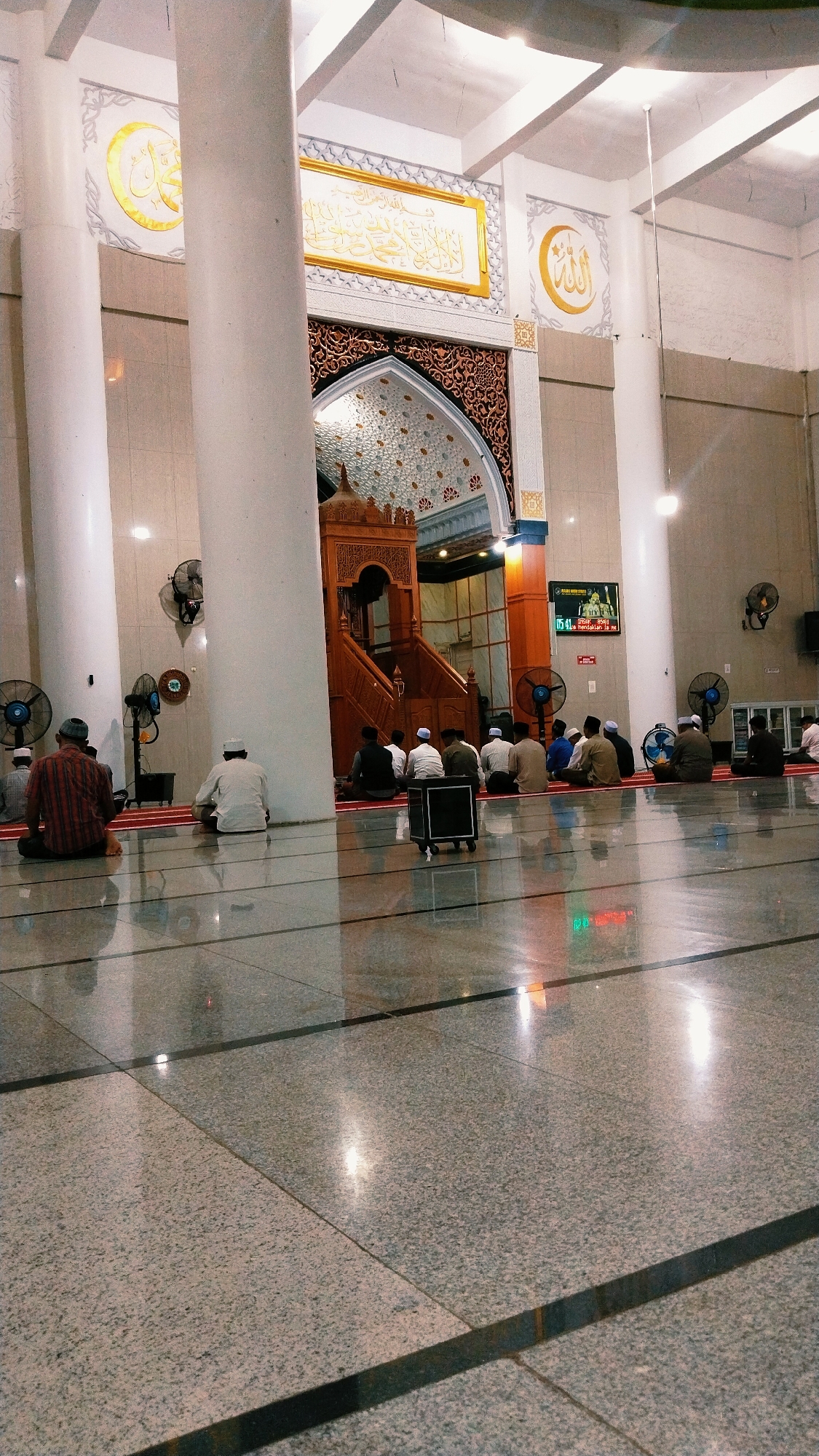 The image depicts the interior of a mosque, presumably in the city of Blangmetimu, Indonesia, with a focus on the prayer area. The scene is dominated by large, white pillars and a polished, reflective floor. In the foreground, several men are seated on red prayer rugs, facing a wooden minbar (pulpit). The men are dressed in traditional Islamic attire, including caps and long shirts or robes. Their posture suggests they are engaged in prayer or meditation.
The background features a decorated archway leading to the minbar, adorned with intricate patterns and calligraphy. Above the archway, large calligraphic inscriptions are visible on the wall. Several electric fans are mounted on the pillars, indicating a warm climate. A digital display screen showing prayer times is also visible on the right side of the mosque. The overall lighting is soft, with reflections from the polished floor adding to the ambiance. The architectural style suggests a modern mosque with traditional Islamic decorative elements.