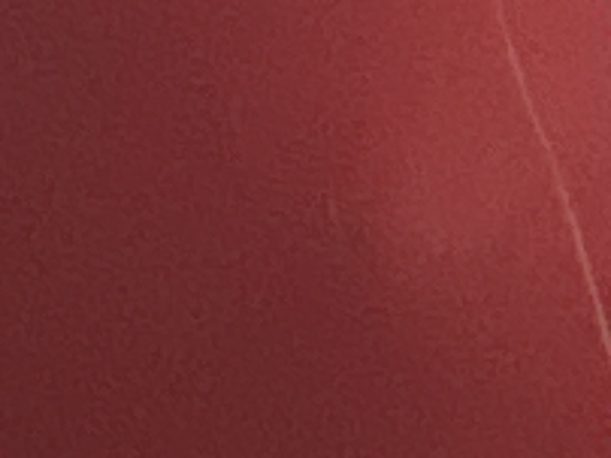 The image is a close-up, abstract view of a red textured surface, possibly fabric or painted material. There is a faint, thin, diagonal line, lighter in color than the surrounding red, that runs from the top right to the bottom left of the frame. The texture appears slightly rough and irregular, with subtle variations in shade and highlights. No other objects, people, or environmental context are discernible.