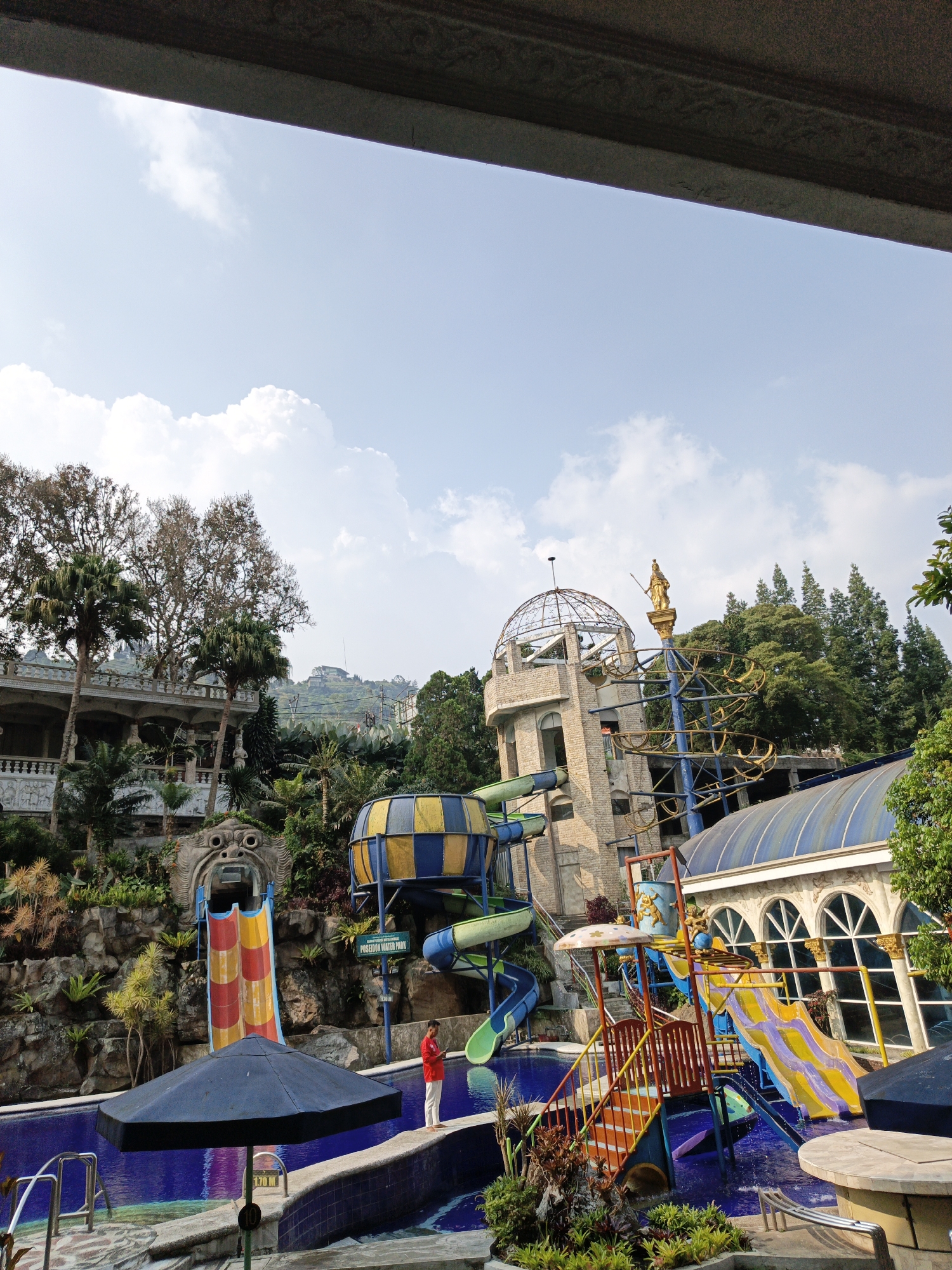 The image depicts a vibrant water park in Cibogo, Indonesia, on a clear day with scattered clouds. Dominating the scene are several water slides, including a large yellow and blue cylindrical slide and a set of multi-colored slides with a playful design. A central tower, adorned with a golden statue at its apex, serves as a prominent architectural feature.
In the foreground, a blue swimming pool reflects the sky and surrounding structures. A person in a red shirt and white pants stands by the edge of the pool, looking at their phone, while a large black umbrella provides shade nearby. Lush greenery, including palm trees and other tropical foliage, is integrated throughout the park, creating an inviting atmosphere. The background features a multi-story building with ornate architectural details and a domed roof, as well as a hill covered in trees. The overall impression is one of a lively and well-maintained recreational area.