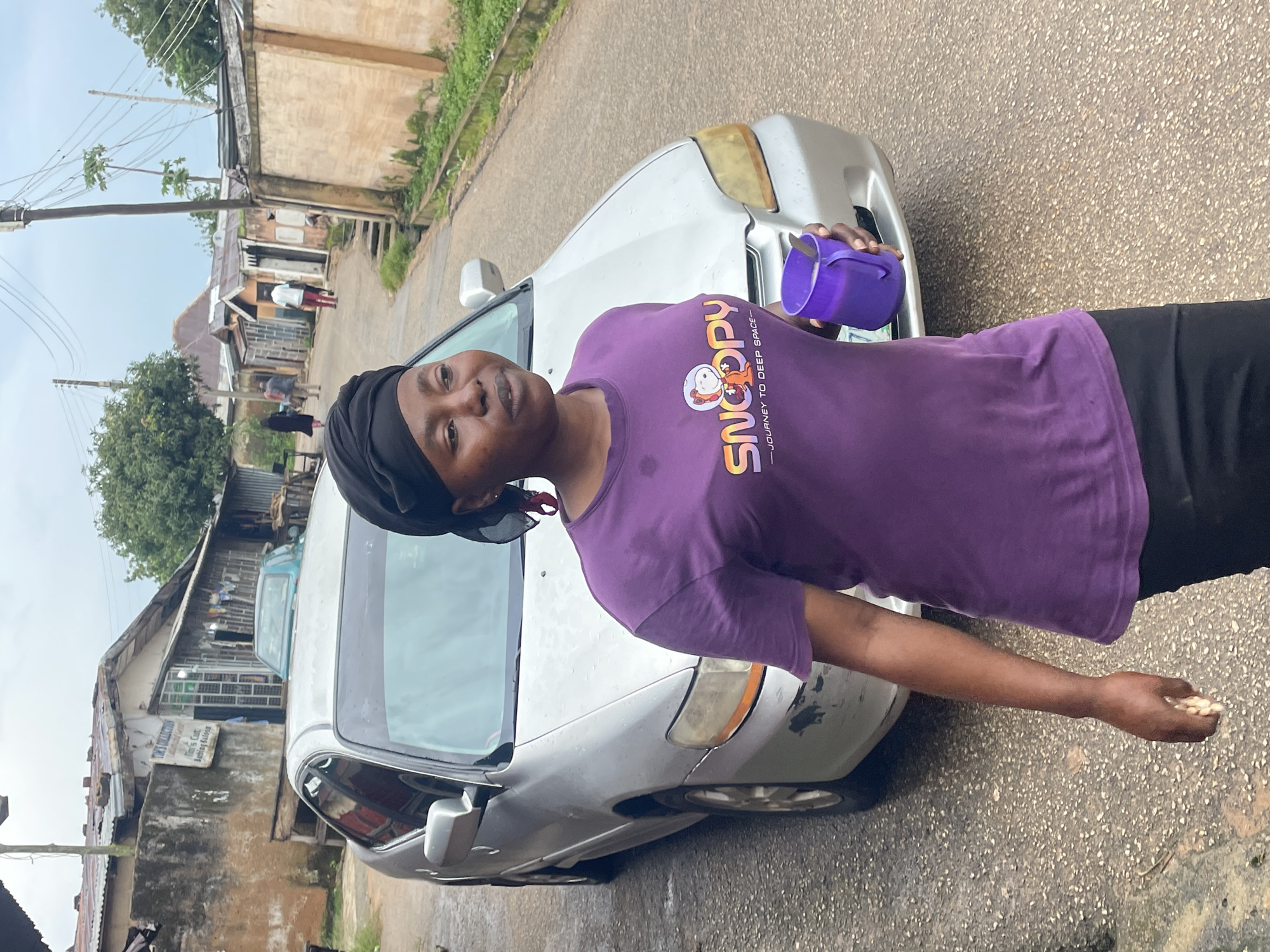 A woman wearing a purple t-shirt with a Snoopy graphic and a black headscarf walks down a paved street in a Nigerian city. She is holding a purple mug with a spoon inside. A silver sedan is parked behind her, and in the background, there are several buildings, some with corrugated metal roofs, and trees. The sky is overcast. The street appears to be a residential or commercial area, likely in Jos, Nigeria, given the architecture and vegetation. The overall scene depicts an ordinary day in a bustling urban environment.