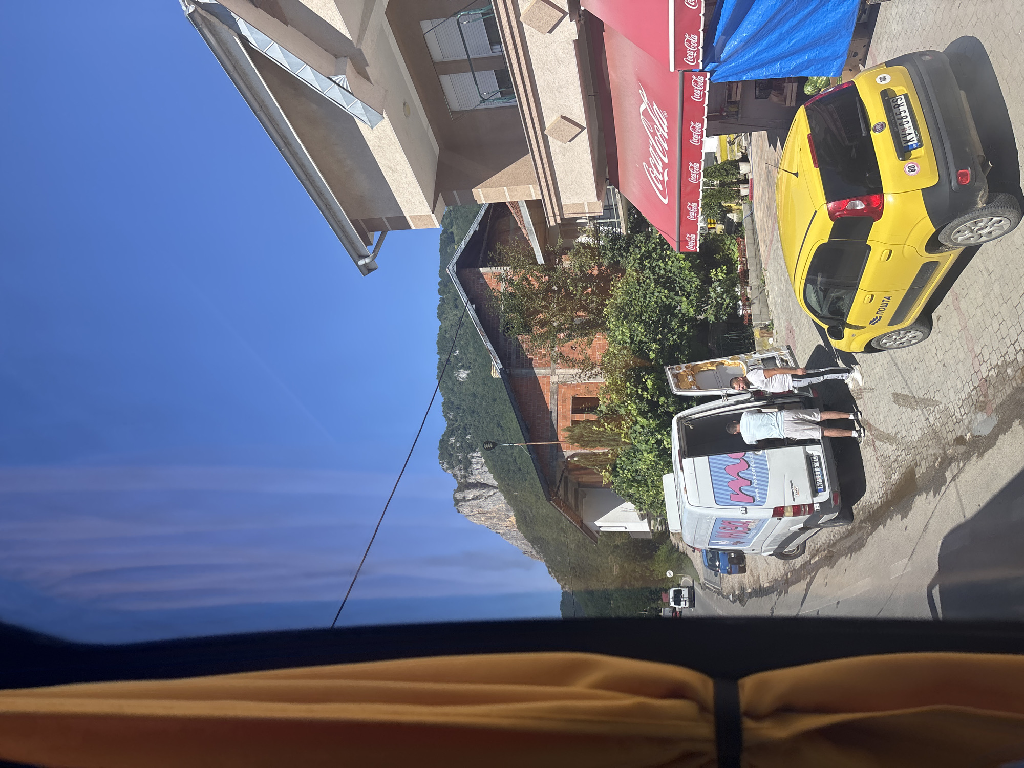 The image, taken from inside a vehicle, captures a street scene on a sunny day. In the foreground, a yellow Fiat Panda, identified as a postal vehicle by "ПОШТА" (Posta) branding, is parked on the right side of the road. It has license plate KV-865-RS. Next to it, a red awning with "Coca-Cola" branding extends over a storefront. A white van with the logo "MSA" and the website "mesacomp.com" is parked on the left side of the road with its rear doors open. Two men are standing near the open van. The road is paved with cobblestones and there are puddles of water. In the background, buildings are visible, including a brick building and a lighter-colored multi-story building. Beyond the buildings, a large, rugged mountain covered in green vegetation dominates the skyline under a clear blue sky. A yellow curtain is visible on the left edge of the frame, indicating the vantage point is from a vehicle with a window.