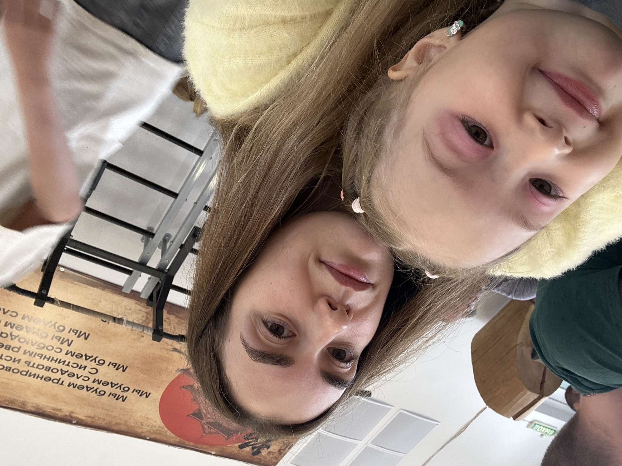 A young woman and a child are in the same space, appearing close together in what seems like a selfie, taken inside a building. The child, who has blonde hair and is wearing a yellow fluffy top, is near the top of the frame, with her face angled towards the camera. The woman has light brown hair and her face is closer to the bottom of the frame. In the background, there is a sign with Russian text including what appears to be "Мы будем тренирово", as well as a grid of lights on the ceiling. Also visible is furniture and what seems to be other people. Based on the text on the sign, it is possible the photo was taken in the city of Chaikovsky, Russia.
