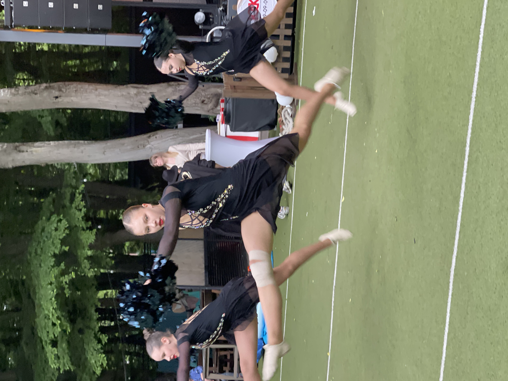 Three young women in black dance costumes with glittering embellishments are performing a synchronized dance routine outdoors on a green, turf-like surface marked with white lines. They are holding black pom-poms. The woman in the center is in mid-leap, with one leg extended forward and the other back. She has a bandage wrapped around her left shin. The woman to her left is also in motion, with her leg extended to the side. The woman to her right is further back and slightly out of focus, also in a dance pose. In the background, there are trees, a speaker system, a white tablecloth-covered table, and some people observing the performance. The scene appears to be an outdoor event or performance. The specific location in Stavropol, Russia, cannot be determined from the image alone.