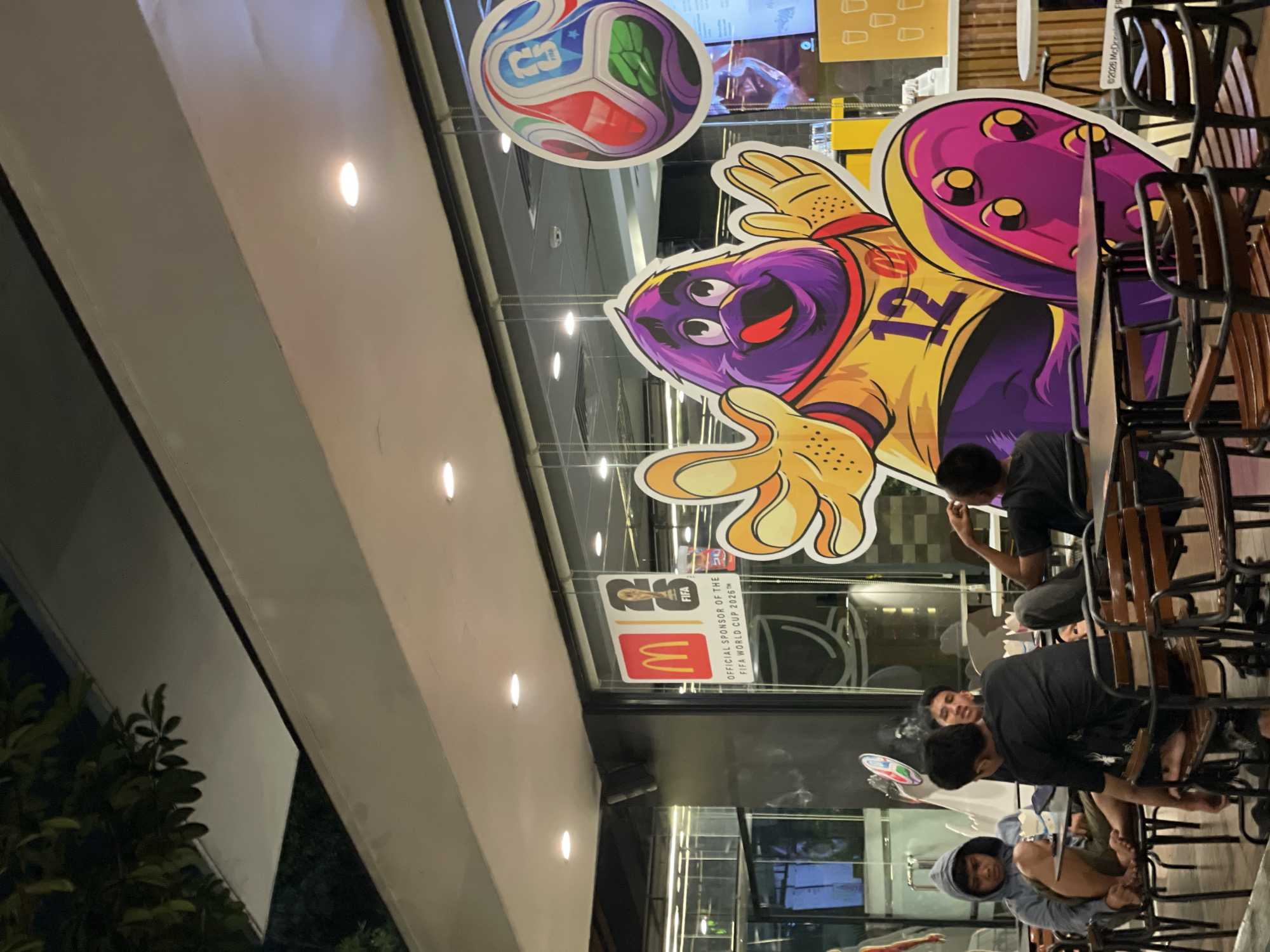 The image captures an outdoor seating area of a McDonald's restaurant at night, located in North Jakarta, Indonesia. In the foreground, several young men are seated at black metal tables and chairs. One man wears a grey hooded sweatshirt and appears to be smoking. Another, wearing a black t-shirt, is also visible, as is a third man with his back to the camera. In the background, a large cardboard cutout of a purple cartoon character wearing a yellow and orange jersey with the number 12 is prominently displayed against the restaurant's glass window. The character is licking its lips. Above the cutout, a colorful soccer ball graphic with FIFA branding is visible. A McDonald's logo and text indicating sponsorship of the FIFA World Cup 2026 are also displayed on the window. The lighting suggests an evening setting, with overhead lights illuminating the scene.
