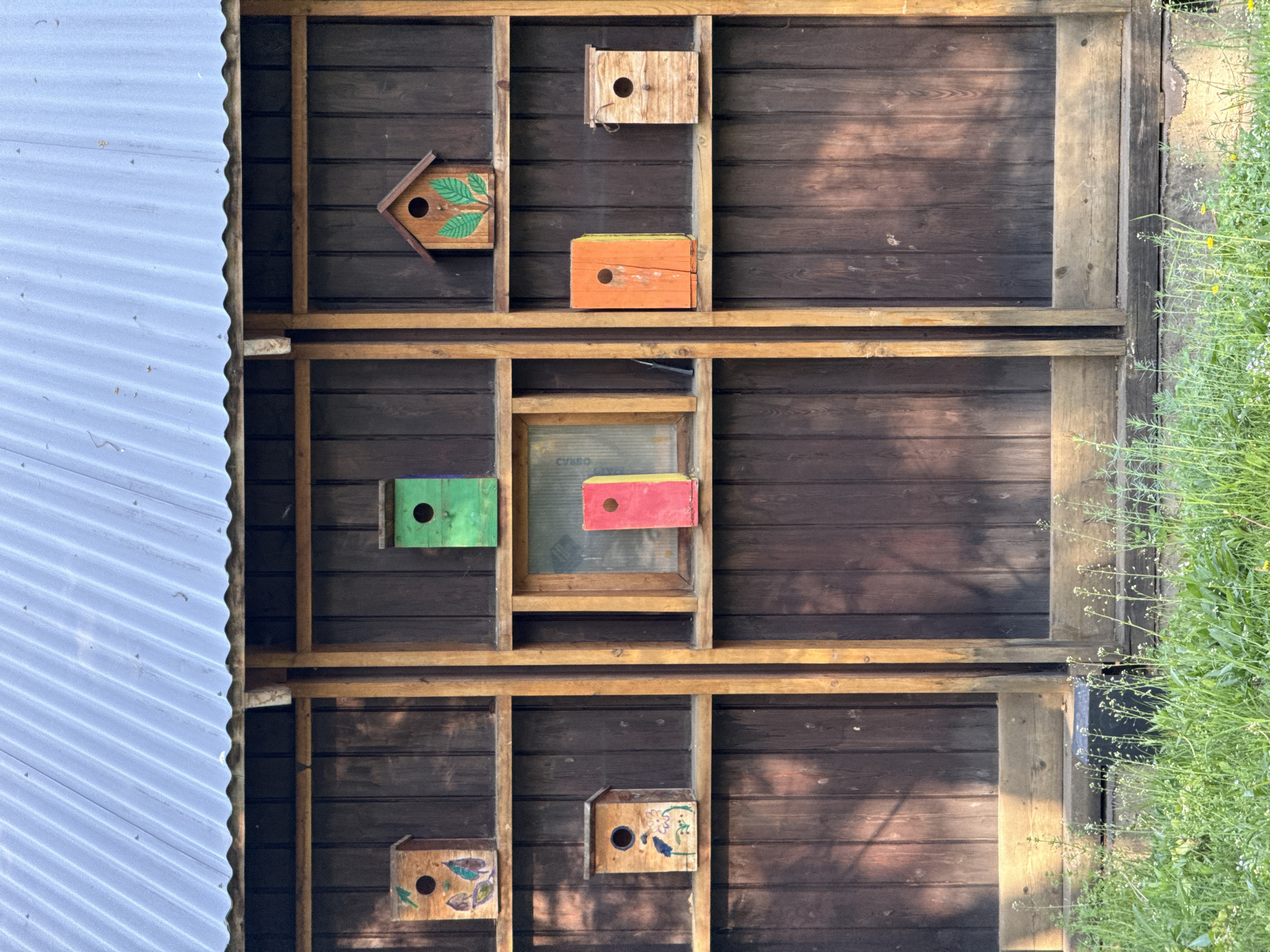 The image shows a wooden structure that appears to be a wall or part of a building with several birdhouses attached to it. The birdhouses are of varying colors and designs. The wooden wall is made of dark brown planks and is divided into sections by light brown wooden supports, creating a grid-like pattern. To the left of the structure is a corrugated metal roof, and on the right side, there is vegetation, possibly weeds or overgrown grass. The image is taken from a slightly low angle, looking upwards. There is no visible indication of the location being Moscow, Russia.