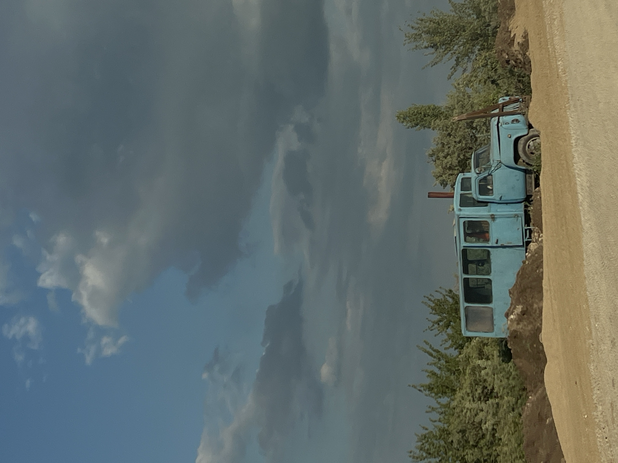 In Khomivka, Ukraine, a light blue, older model truck with a bus-like rear compartment is parked on a dirt road, partially obscured by mounds of dirt and sand. The vehicle features large windows along its side and a driver's cab with a visible steering wheel. The environment is rural, with a line of green trees bordering the background and a vast sky filled with dramatic, dark storm clouds interspersed with patches of blue. The road surface appears rough and uneven. There are no people visible in the image.