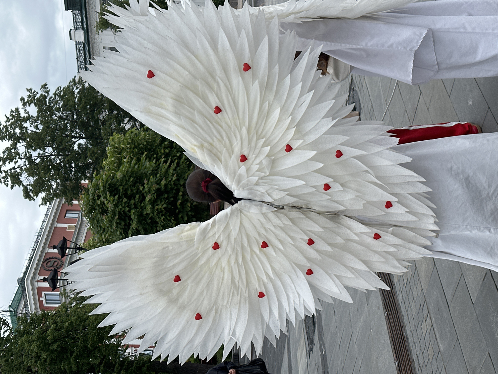 A person wearing large white feathered wings adorned with small red hearts stands on a gray brick pavement. The wings span a significant width, constructed from layers of white material that resembles feathers. The person is wearing a white garment. A red and white stripe is visible below the white garment.
In the background, lush green trees and foliage provide a natural backdrop. Buildings with red brick facades and ornate architectural details can be seen behind the trees. The street lamps in the background are ornate with globe lights on top of each lamp. A cloudy sky appears overhead.
The location is the city of Nizhny Novgorod, Russia, based on the architectural style of the building in the background.