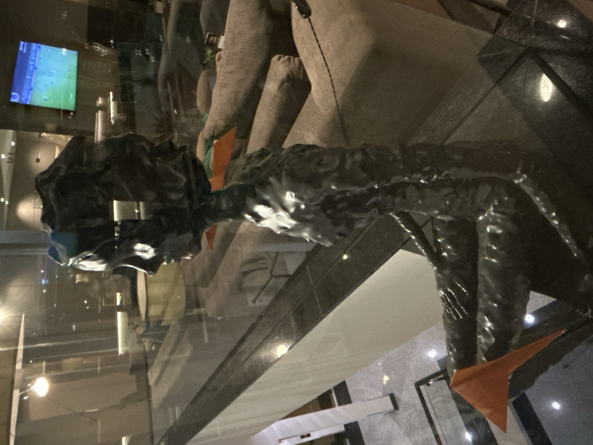 A sculpture of a seated figure with dark, textured skin and an orange triangular object resting on its lap is positioned on a glass railing overlooking a lower level of a building. The sculpture is in profile view, facing left, and appears to be made of a metallic material. The figure's head is adorned with a dark, textured headdress. In the background, a television screen displays a soccer match, and plush, brown seating is visible. The environment appears to be an indoor space, possibly a lobby or lounge, with modern architecture and lighting. The image is taken from a high angle, looking down and slightly to the right.