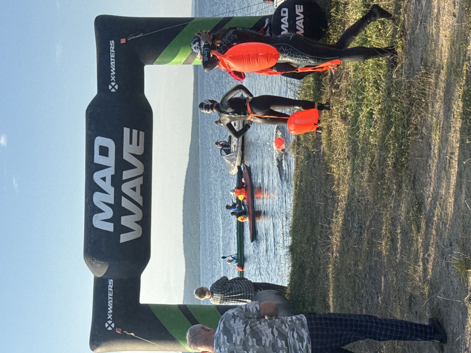 An inflatable archway with "MAD WAVE" branding stands at the edge of a body of water. Three people in full-body wetsuits are near the archway, two of them facing away from the camera. One person is wearing a black wetsuit with an orange float attached to their waist. Another person is wearing a patterned black wetsuit, also with an orange float. The third person, also in a wetsuit, is facing the water. In the background, several kayaks and a small boat are on the water. The landscape behind the water consists of rolling hills. The ground in the foreground is covered with dry grass and dirt. The sky is clear and blue.