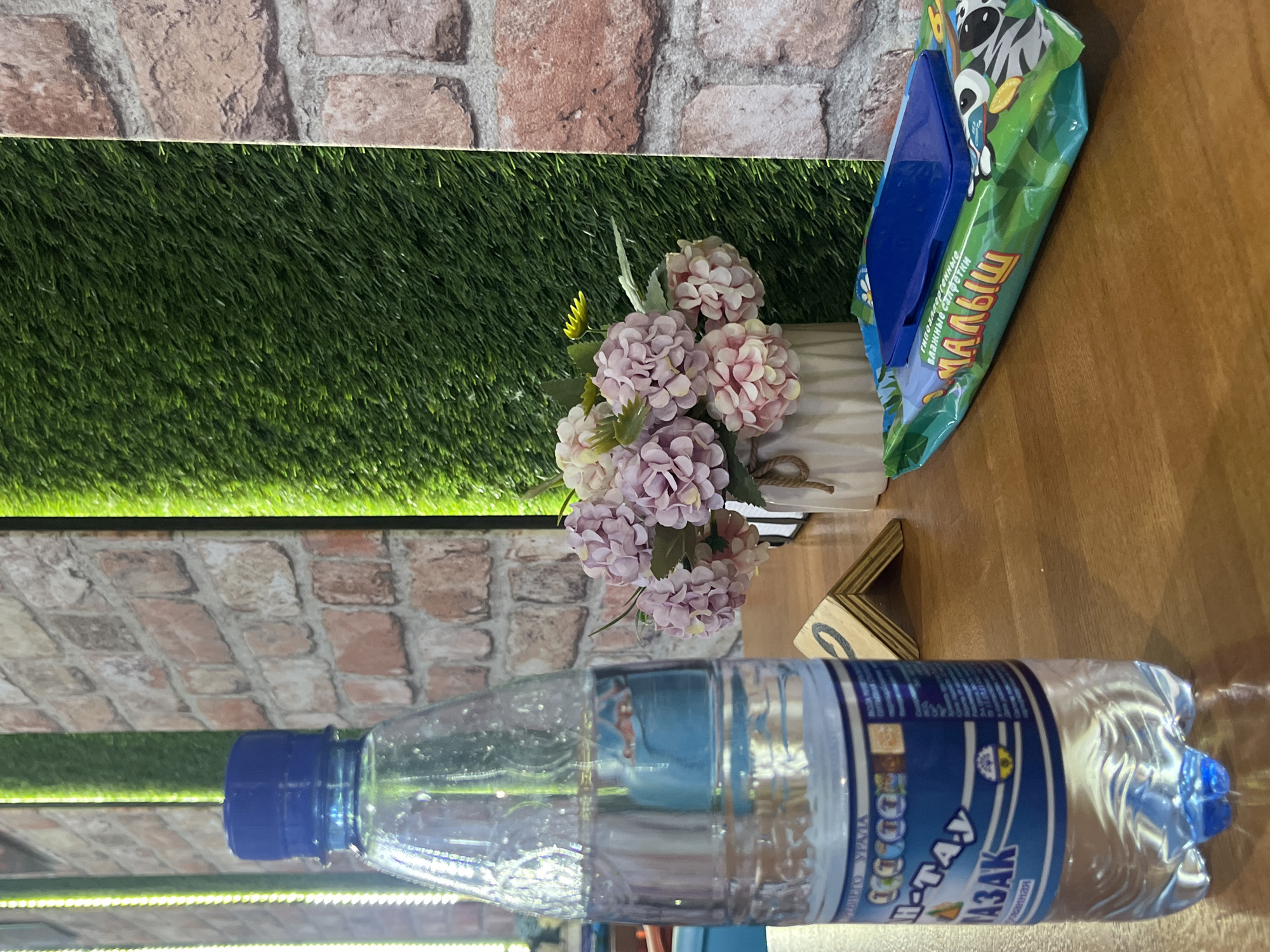 The image captures a still life arrangement on a wooden surface, set against a backdrop of artificial brick and grass. In the foreground, a clear plastic bottle of water lies on its side, its blue cap visible. The bottle appears to be partially filled with water, with some condensation visible on its surface. The label on the bottle is predominantly blue and features text in what appears to be Cyrillic script, including "Н-ТАУ" and "УРАЛА".
Behind the water bottle, a small bouquet of artificial pink and white flowers is arranged in a white vase, which is partially obscured by the flowers. To the right of the flowers, a package of baby wipes is displayed. The packaging is brightly colored green and features cartoon illustrations of zebras and other animals, along with Cyrillic text, notably "МАЛЫШ".
The background consists of a textured wall that mimics red brick, situated above a strip of artificial green grass. The lighting in the image suggests an indoor setting, possibly a table in a cafe or home. There are no people visible in the frame. The overall impression is of a simple, decorative arrangement. The specific location is identified as Chelyabinsk, Russia, based on the bottle label's likely reference to "Uralataw" water, a brand originating from the Ural Mountains region, which includes Chelyabinsk. The time of day and weather are not discernible from the image.