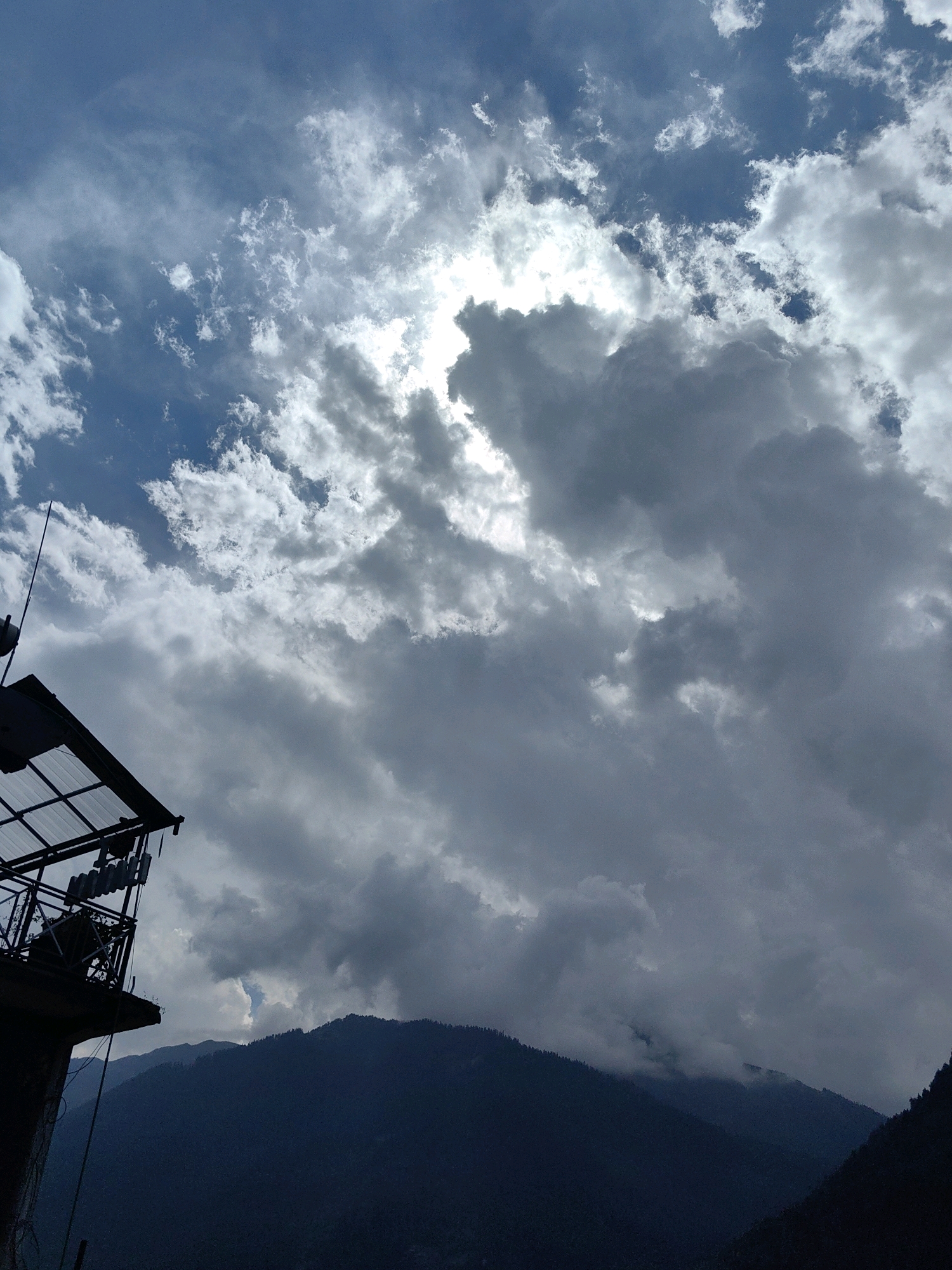 A silhouette of a structure with a metal roof and what appears to be telecommunication equipment is visible in the lower left corner of the image. The structure is dark and stark against a dramatic sky filled with a mix of dark, stormy clouds and lighter, brighter clouds where the sun is partially obscured. Below the sky and clouds, a dark, heavily forested mountain range forms the background. The overall scene suggests a mountainous region, potentially Vashisht, India, under a dynamic sky. There are no people visible in the image.