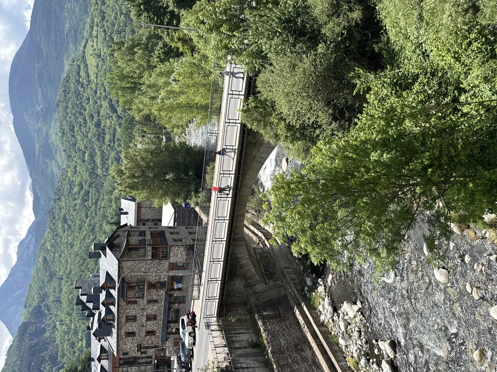 The image shows a view in Benasque, Spain, looking down at a stone bridge spanning a river. People are walking on the bridge. The river flows from left to right with large rocks scattered in the water. On the left of the bridge is a street with cars parked and a stone building with multiple windows and a slate roof. Behind the building is a mountain covered in trees. The sky is visible with some clouds. Trees line both sides of the river. The image appears to be taken from an elevated position.