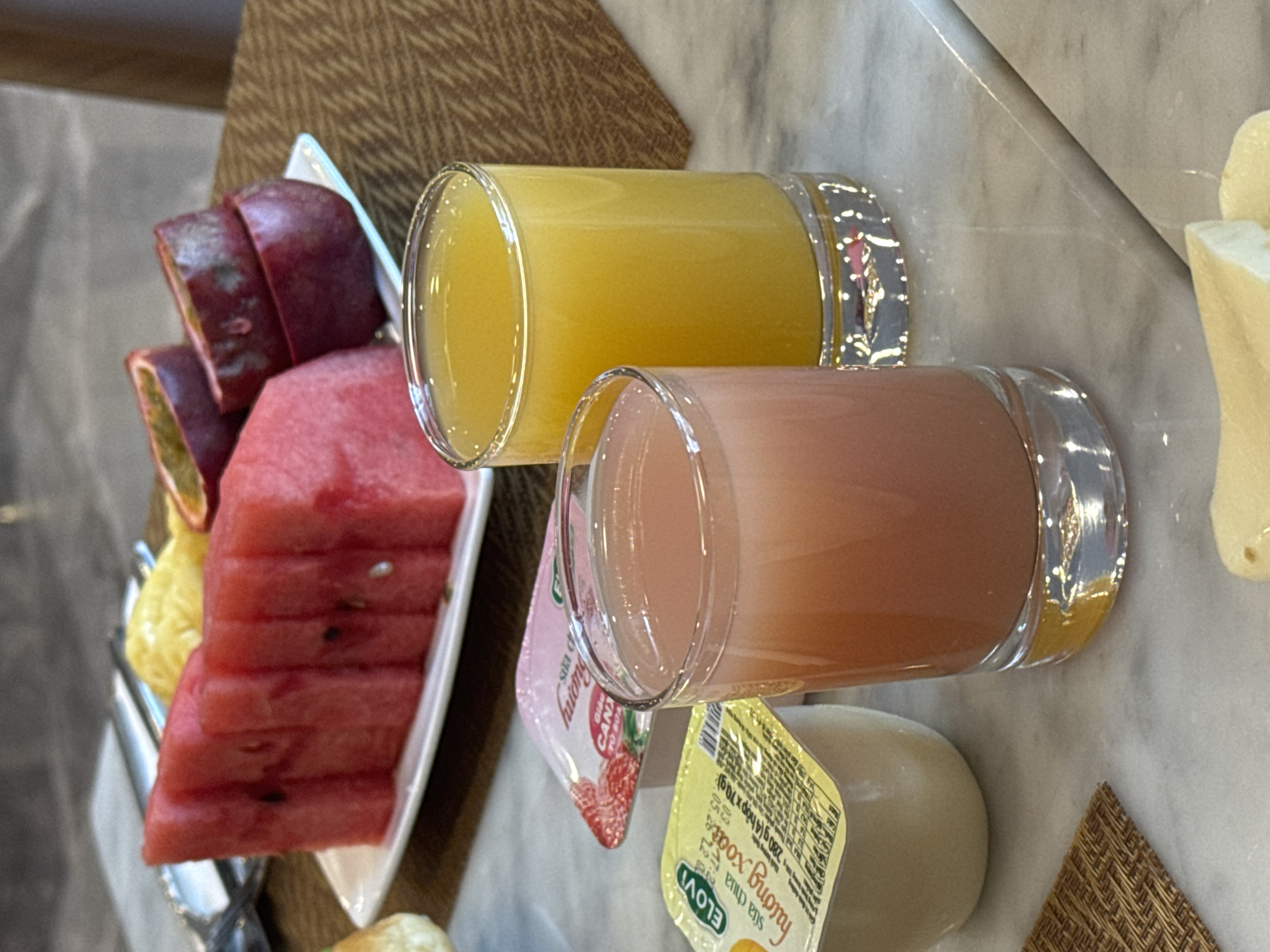 The image shows a selection of breakfast items arranged on a marble surface, seemingly indoors. On the left, a white rectangular plate contains a stack of sliced watermelon, passion fruit, and yellow fruit (possibly pineapple or mango). To the right of the plate, two glasses are placed side by side, one filled with orange juice and the other with a pink juice, likely grapefruit. Next to the pink juice, there are two small containers of yogurt, one with a strawberry flavor and the other with a yellow label written in Vietnamese script that includes the words "ELOVI" and "sữa chua hương xoate," indicating yogurt with an unspecified flavor. A piece of cheese is visible on the right edge of the frame. The scene suggests a hotel breakfast setting, possibly in Đà Nẵng, Vietnam, given the Vietnamese yogurt brand. The focus is on the food and beverage arrangement, with no people visible in the frame.