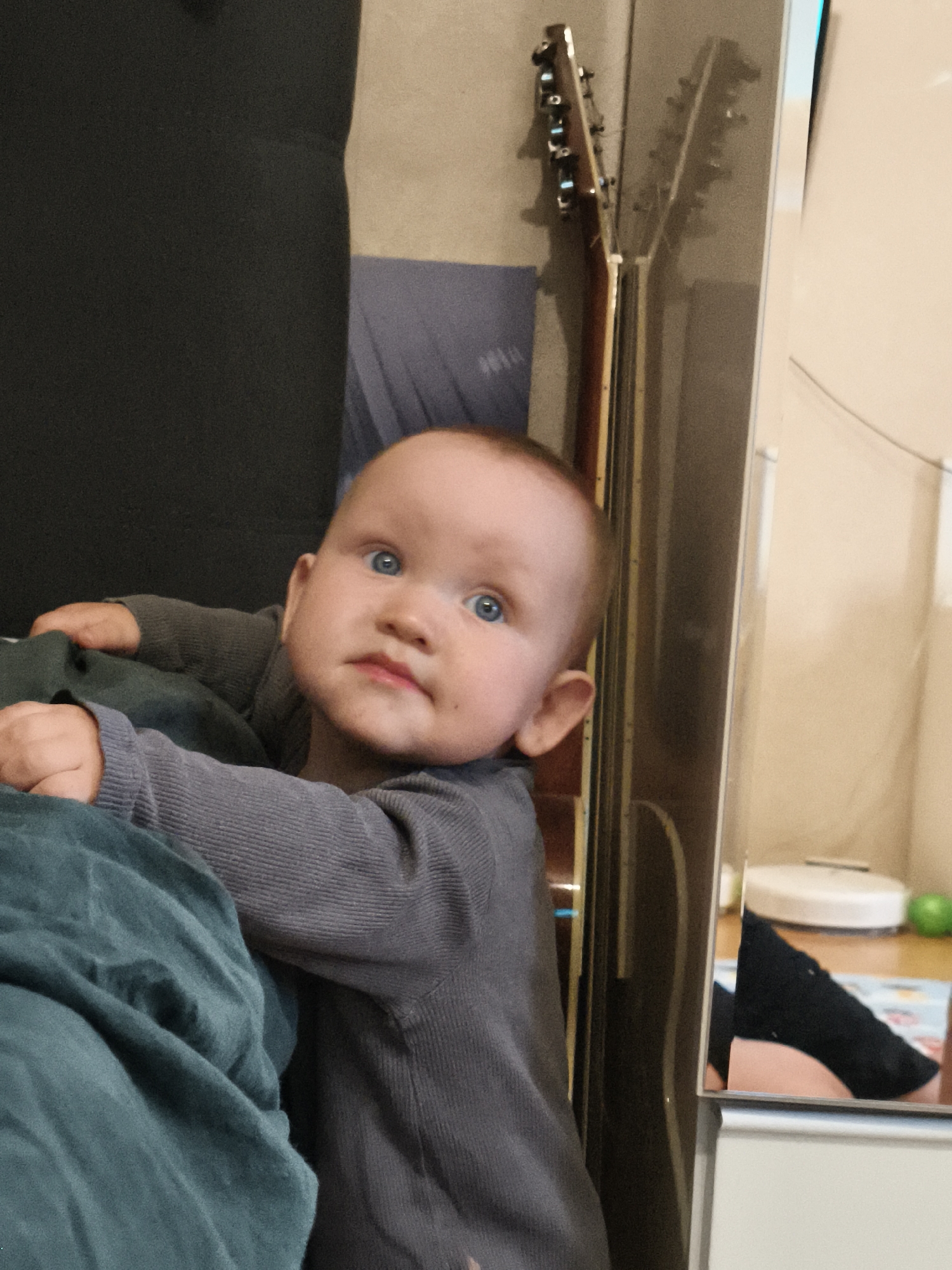 A baby with blue eyes and light skin is lying on a teal blanket, leaning on a dark grey object. The baby is wearing a dark grey long-sleeved shirt. Behind the baby, a guitar is leaning against a wall. To the right of the baby, a mirror reflects a person's legs wearing black socks and a pink object on a table. The setting appears to be a home in Chaikovsky, Russia.
