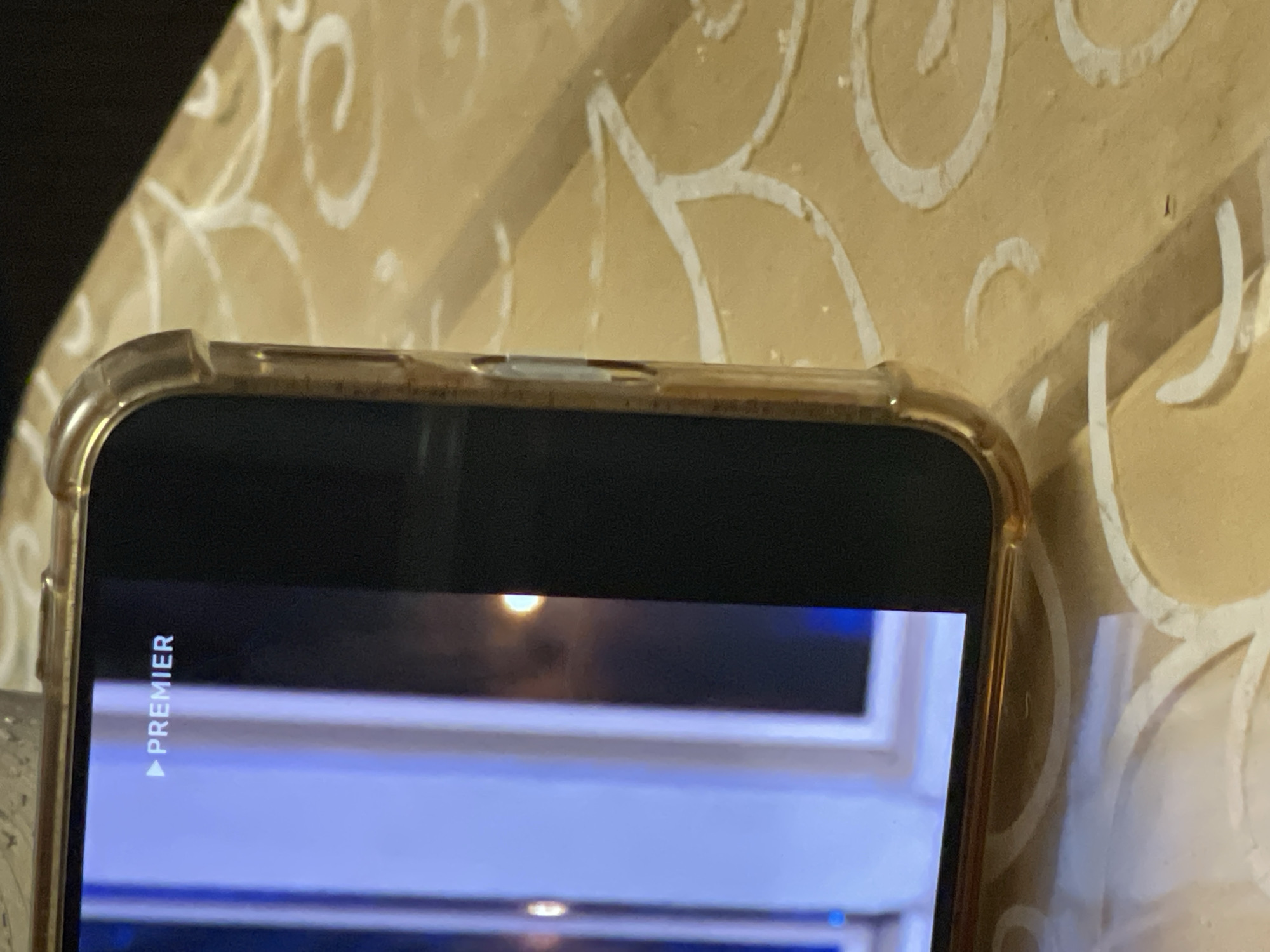 A close-up, side-view photograph shows a smartphone screen displaying the word "PREMIER" with a play icon to its left, superimposed on a slightly blurred background. The phone is positioned on a table with a light yellow textured surface featuring white swirling patterns. The phone itself has a gold-colored edge and a clear protective case. The lighting in the image suggests an indoor setting, possibly at night, given the dim reflections on the screen and the overall ambiance. The scene does not provide any specific geographical markers to indicate its location within the city of Mirnyy, Russia.