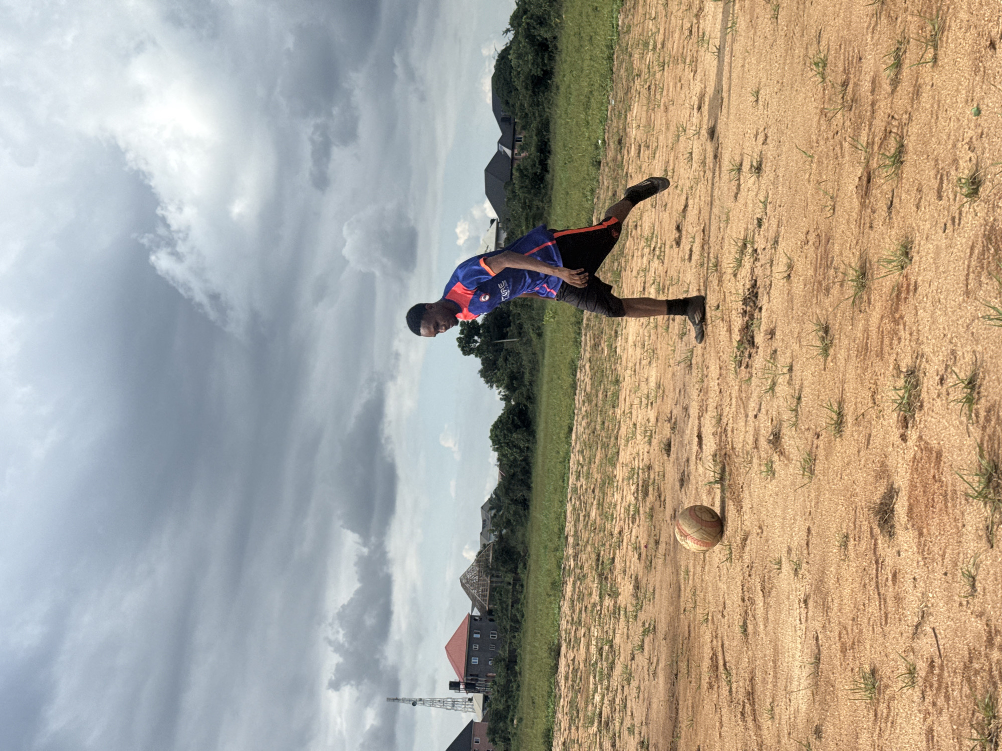 A young man in a blue and red soccer jersey and black shorts is playing soccer on a sandy, sparsely vegetated field. He is in mid-stride, with his left leg extended as if kicking a ball. A soccer ball rests on the ground to his left. In the background, there is a line of green trees, houses under construction, and a cellular tower against a cloudy sky. The scene is set in Ihiagwa, Nigeria.