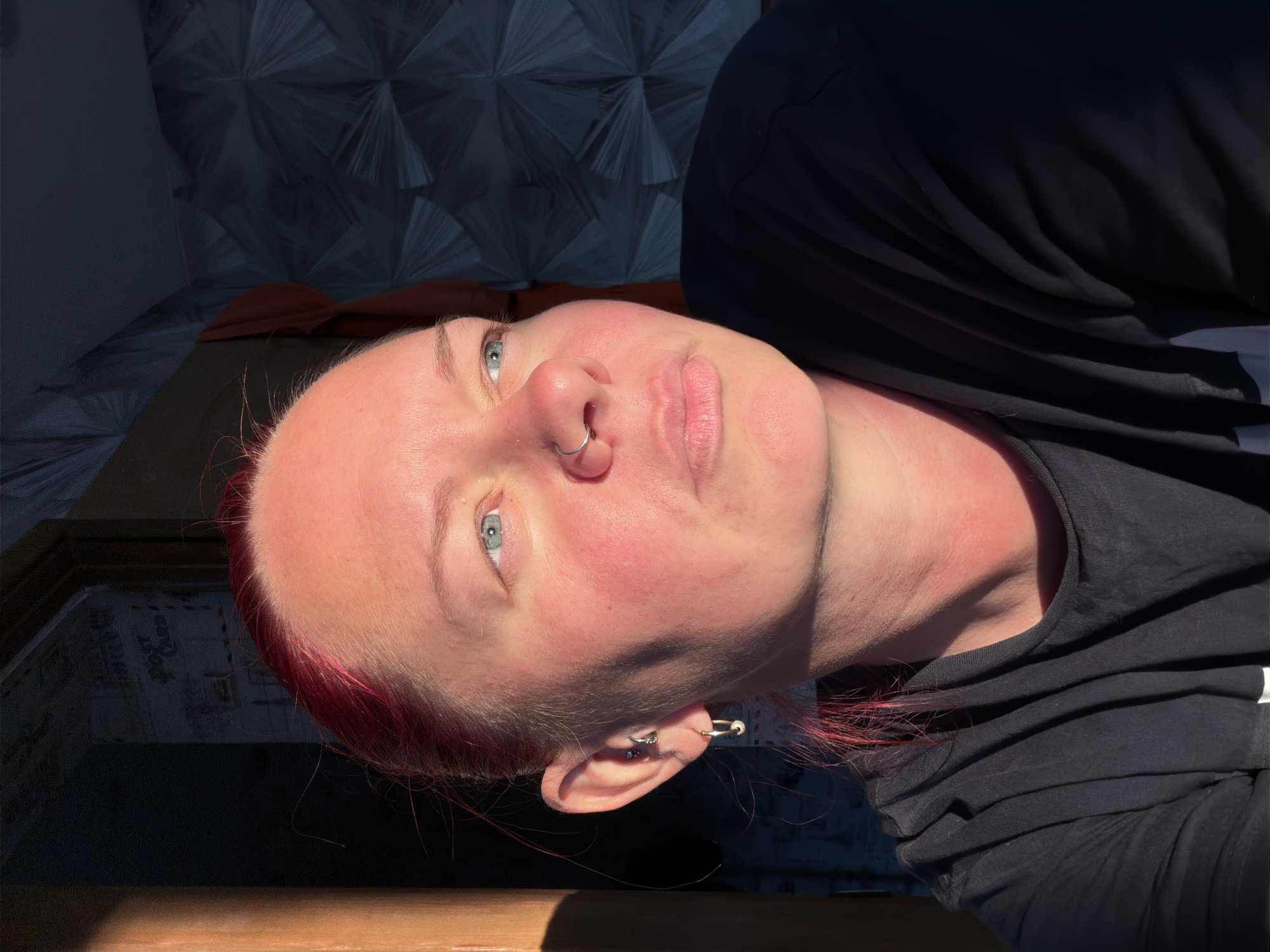 The image shows a close-up portrait of a person with bright pink hair, facing to the right with their gaze directed upwards and to the right. The person is wearing a black t-shirt and has a nose ring. Their skin is flushed, likely due to warm lighting. The background features a dark, patterned wallpaper. A vertical wooden object is visible on the left edge of the frame, casting a shadow. The scene appears to be indoors, with natural light streaming in from an unseen source, creating a strong contrast between light and shadow. The overall impression is one of a candid, personal moment captured in a domestic setting. The location is identified as Chaikovsky, Russia.