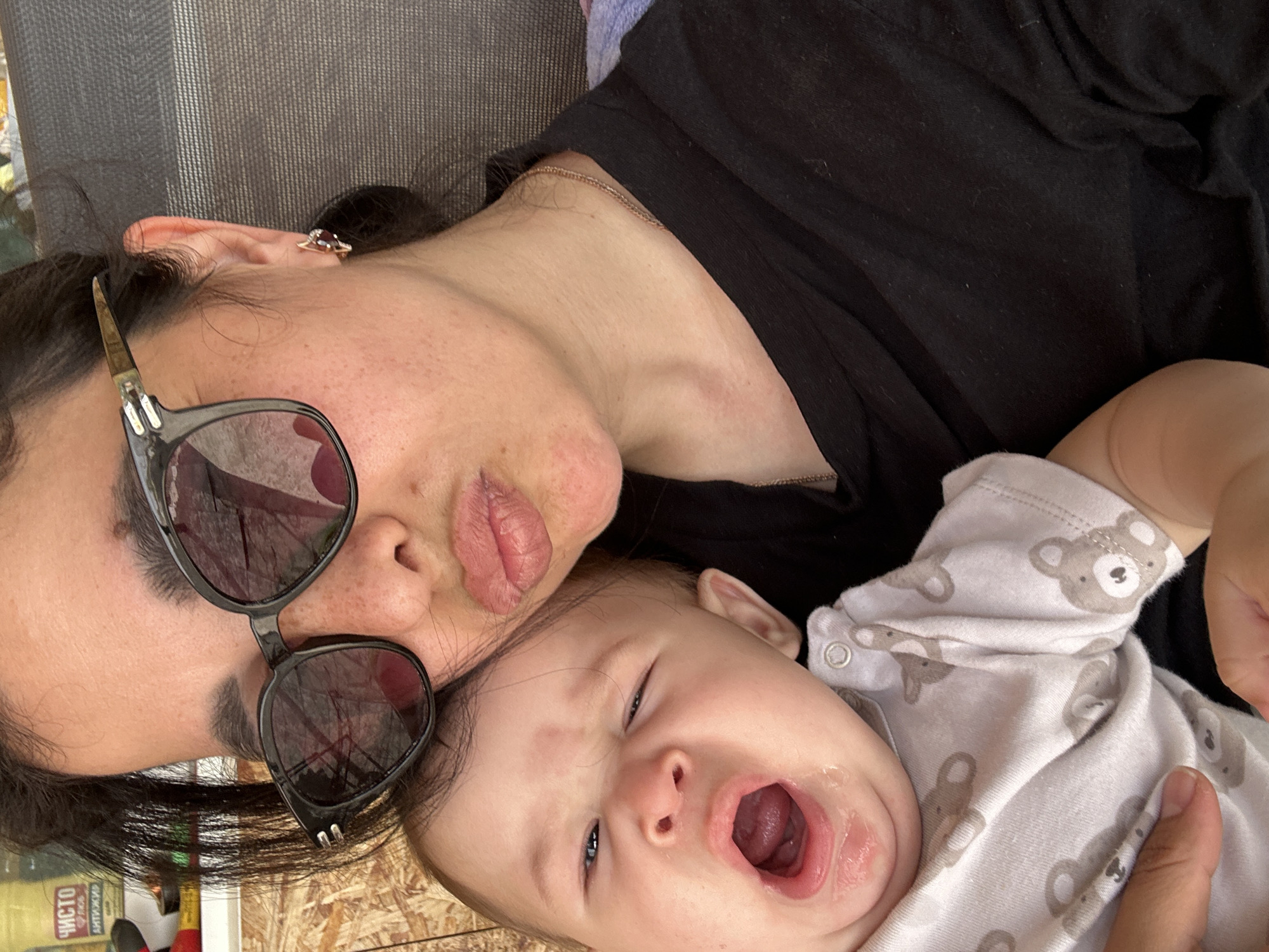 A woman wearing black sunglasses and a black shirt lies on a wooden surface next to an infant wearing a white onesie with bear designs. The infant is yawning with its mouth wide open. The woman is wearing an earring on her left ear. A bottle of cleaning solution is visible in the lower left corner of the image. The location is Unknown, Unknown.