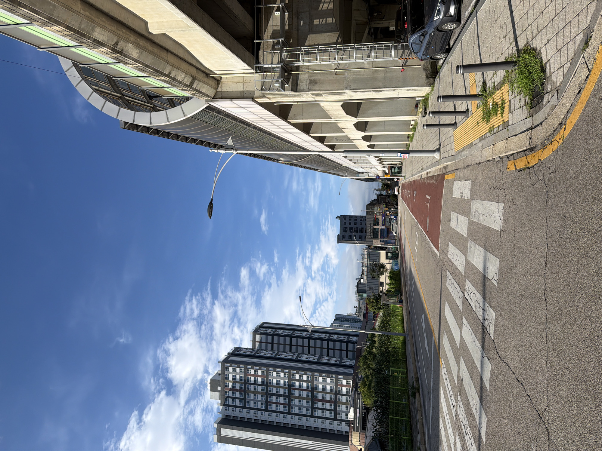 This is a street-level, eye-level, medium shot taken on a sunny day in Dongducheon-si, South Korea. The image captures a paved road with a marked crosswalk in the foreground. To the right of the road, there is a sidewalk with tactile paving, bollards, and overgrown grass. Beyond the sidewalk, a multi-story building with arched openings and a light-colored facade extends upwards, partially obscured by an elevated structure with a curved, metallic roof and green-tinted windows. A street lamp is visible on the left side of the road, and in the distance, tall, modern apartment buildings stand against a blue sky with scattered white clouds. A dark-colored sedan is parked on the right side of the road near the sidewalk. The road itself has a red-painted section and white line markings.