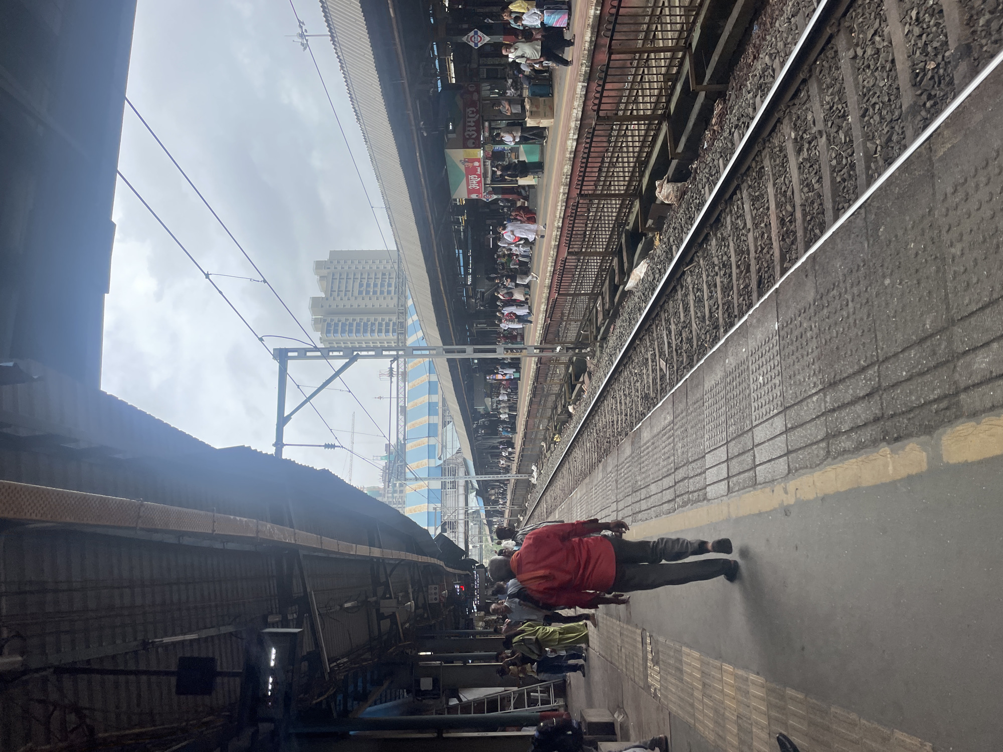 This image depicts a busy Mumbai suburban railway station on an overcast day. The perspective is from a platform looking down a set of train tracks, with another platform and its roof visible across the tracks.
On the foreground platform, a man in a red shirt and dark pants walks away from the camera, towards the right. Several other people are visible on the platform, some standing, some walking. There are a few ladders leaning against a support structure on the left side. The platform itself is paved with textured tiles and bordered by a yellow line.
Across the train tracks, the second platform is lined with people waiting. Various stalls and signs are visible on this platform, including one with "अमूल" (Amul) written on it, indicating a commercial presence. The platform is covered by a long, sloped roof supported by pillars, and beyond that, a modern multi-story building stands against the cloudy sky. Overhead, electrical wires for the trains are strung between poles. The train tracks are filled with ballast. The overall scene suggests a typical, bustling day at a Mumbai railway station.