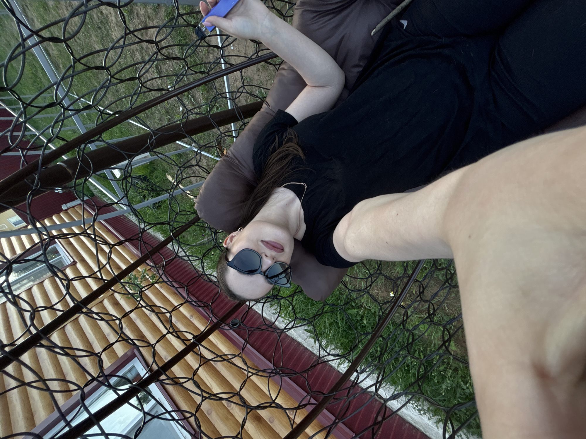 A young person is depicted relaxing outdoors, lying in a black metal hanging chair or hammock, captured from a low, upward-looking perspective. They are wearing dark sunglasses, dark lipstick, and a black top, with a thin gold chain visible around their neck. Their long dark hair cascades over their shoulder. They rest on a brown cushion and are holding a purple electronic device, possibly a vape pen, in their left hand. The person's posture suggests a moment of leisure.
The background reveals a rustic building with light-colored, horizontal wooden siding and red trim along the roofline, indicative of a dacha or rural home, consistent with the Rusalevka, Russia location context. Two windows are visible on the building's facade. Beyond the structures, green grass and sparse vegetation are visible through the intricate mesh pattern of the hanging chair. The lighting suggests it is daytime, likely a pleasant late afternoon or early evening, with clear weather. No visible text is present in the image.