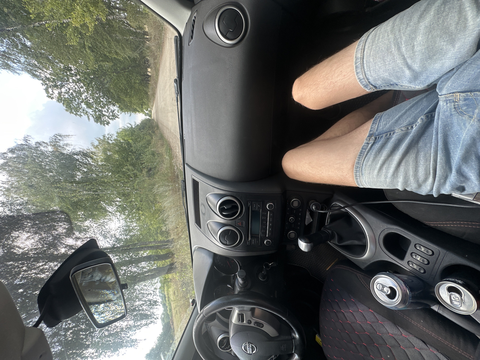 An individual's legs, clad in denim shorts, are positioned prominently in the foreground of the image, resting within the passenger cabin of a car. The car's interior features a black dashboard with air vents, a central console with a gear shifter and what appears to be a radio or media player. Two open beverage cans are visible in the cup holders. The view through the windshield reveals a dirt road stretching into the distance, flanked by trees and foliage, under a partly cloudy sky. The scene suggests a casual drive through a rural or semi-rural environment. The image may have been taken in or near Tynna, Ukraine, given the context provided.