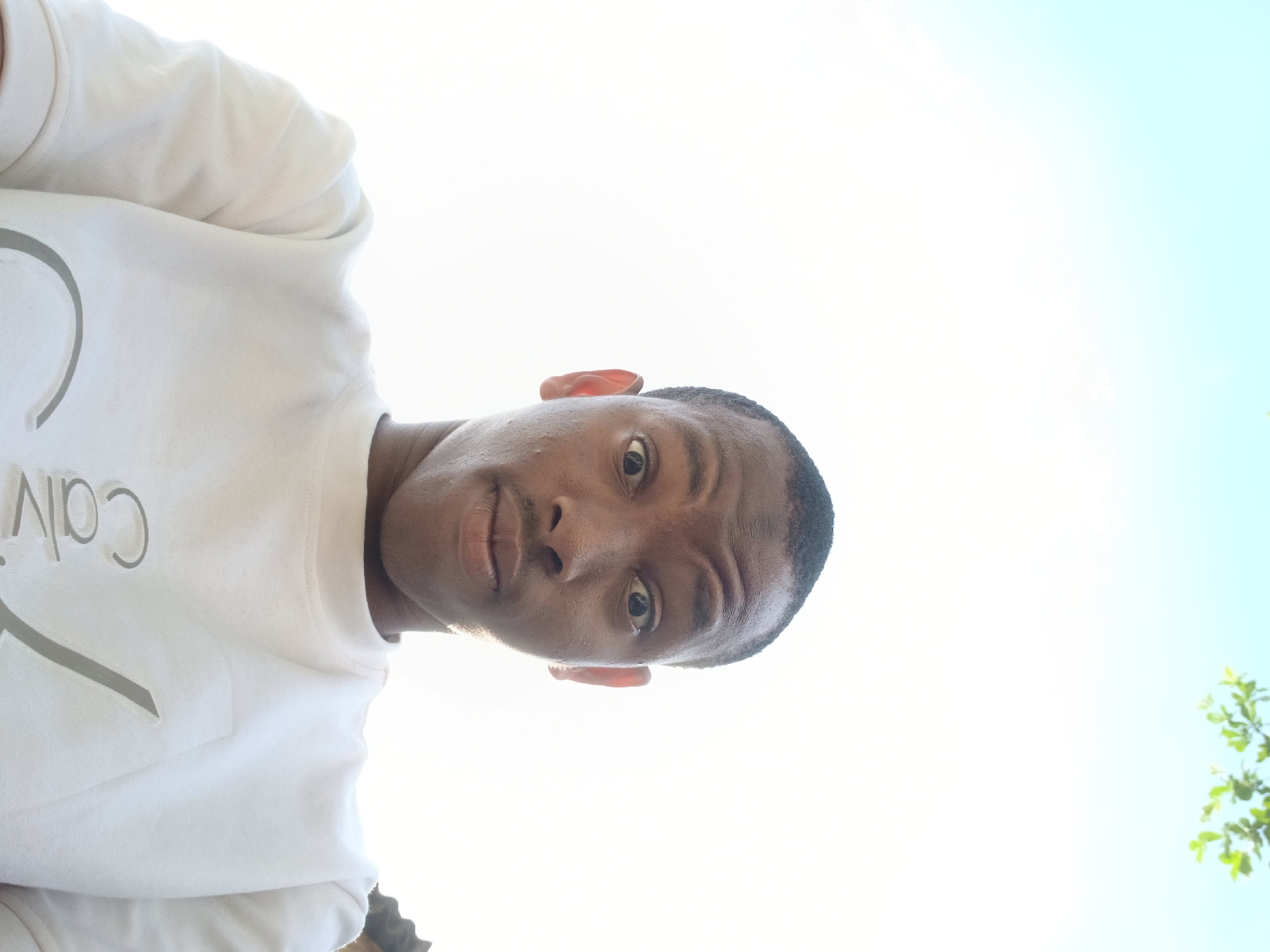 An eye-level photograph shows a dark-skinned man from a low angle. He has short, dark hair. The man is wearing a white, long-sleeved sweatshirt with "Calvin" visible on the front. His face is in focus, and the bright light washes out some of the details. The background is a light blue sky with some green leaves in the corner. The location, Abule Ijesa, Nigeria, cannot be verified from the visual content of the image.