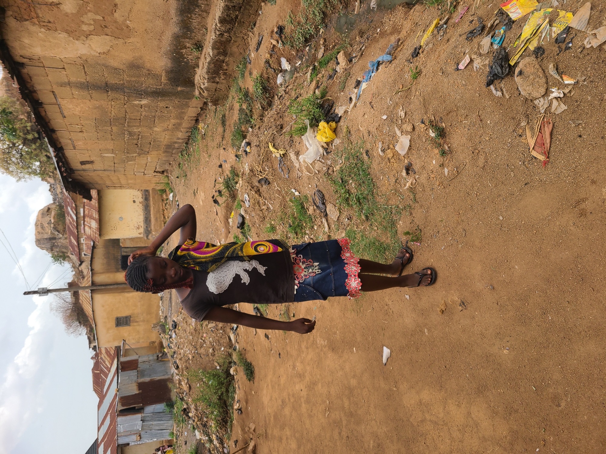 A young woman stands in a littered, sandy area of Jos, Nigeria. She wears a dark t-shirt with a light-colored design, a dark denim skirt with red and white trim, and dark sandals. A colorful textile is draped over her shoulder. Her hair is braided. The surroundings include buildings with corrugated metal roofs and brick or mud walls. Trash and vegetation are scattered around the sandy ground. The sky is overcast.