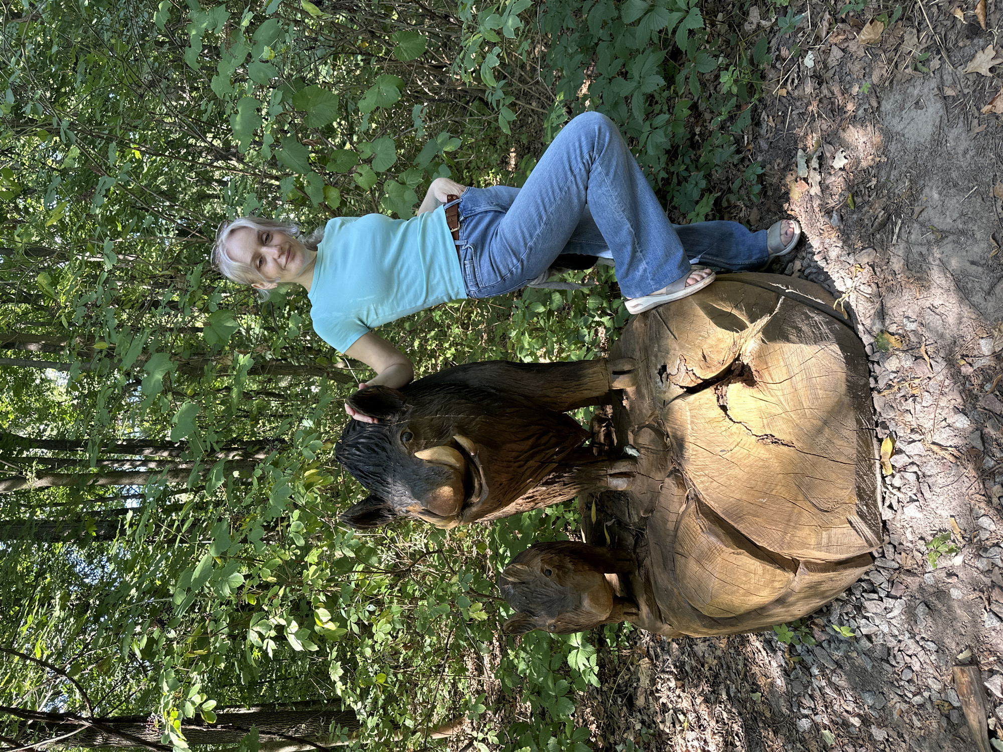 A young woman with blonde hair pulled back in a ponytail poses for a photo in a forest. She is wearing a teal t-shirt, blue jeans, and tan sandals. The woman is standing to the right of the image, leaning with her left hand on a large wooden sculpture of a wild boar, with a smaller wild boar at its feet. The sculpture is resting on a large, rough-hewn log. The background is a dense forest with green foliage and tall trees, suggesting a wooded area. The ground is covered with leaves, dirt, and small stones. The lighting indicates it is daytime, with sunlight filtering through the trees. The location is identified as the "Park of Partisan Glory" in Kyiv, Ukraine.