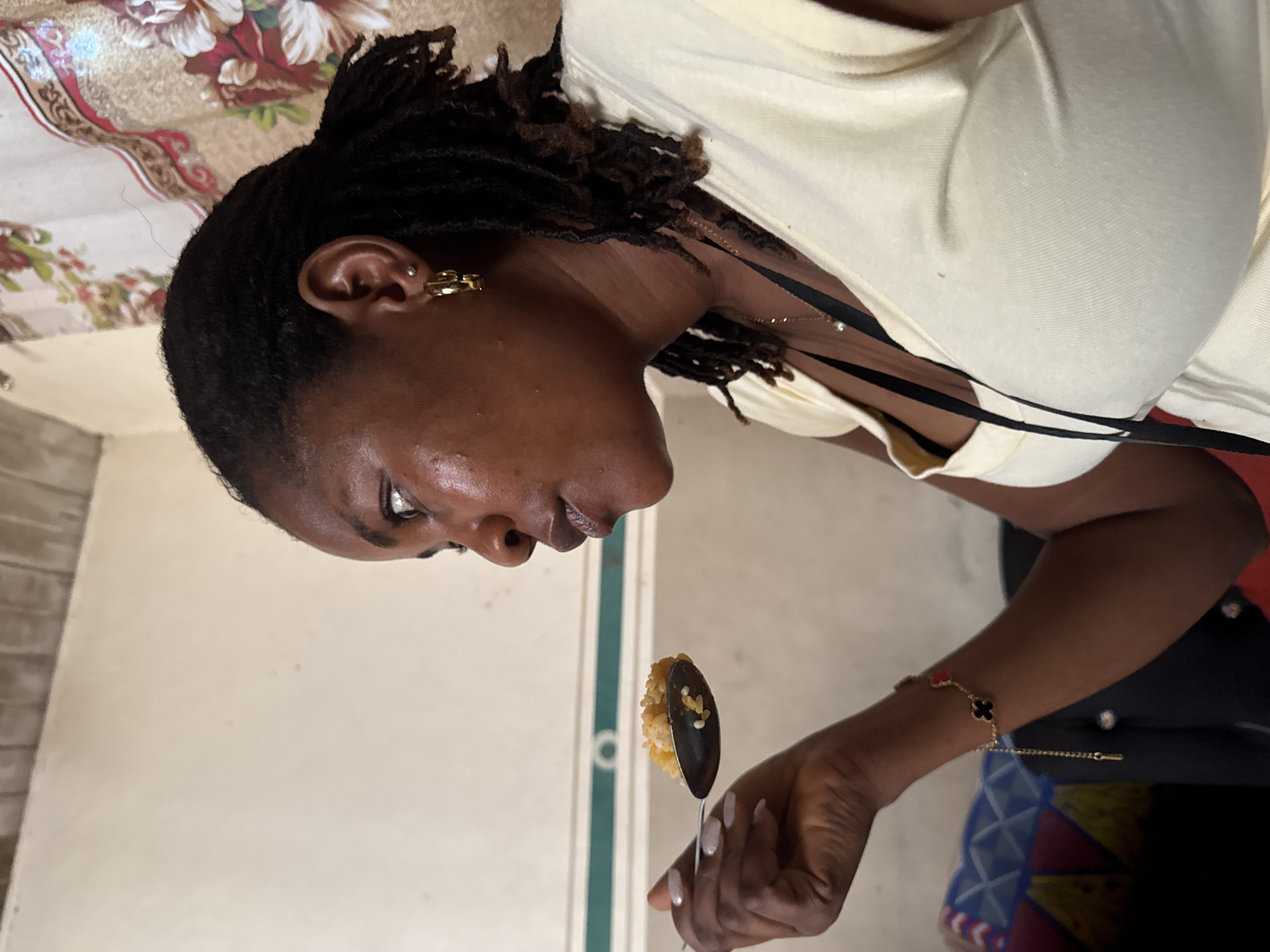 A woman is looking at and holding a spoonful of food. She has dark hair in locs and is wearing gold earrings and a light yellow shirt. She has a gold bracelet on her wrist. The background includes a wall and part of a floral patterned curtain. There is also a green and white vertical line, likely a doorframe or wall detail. The location of the image is Badagry, Nigeria.