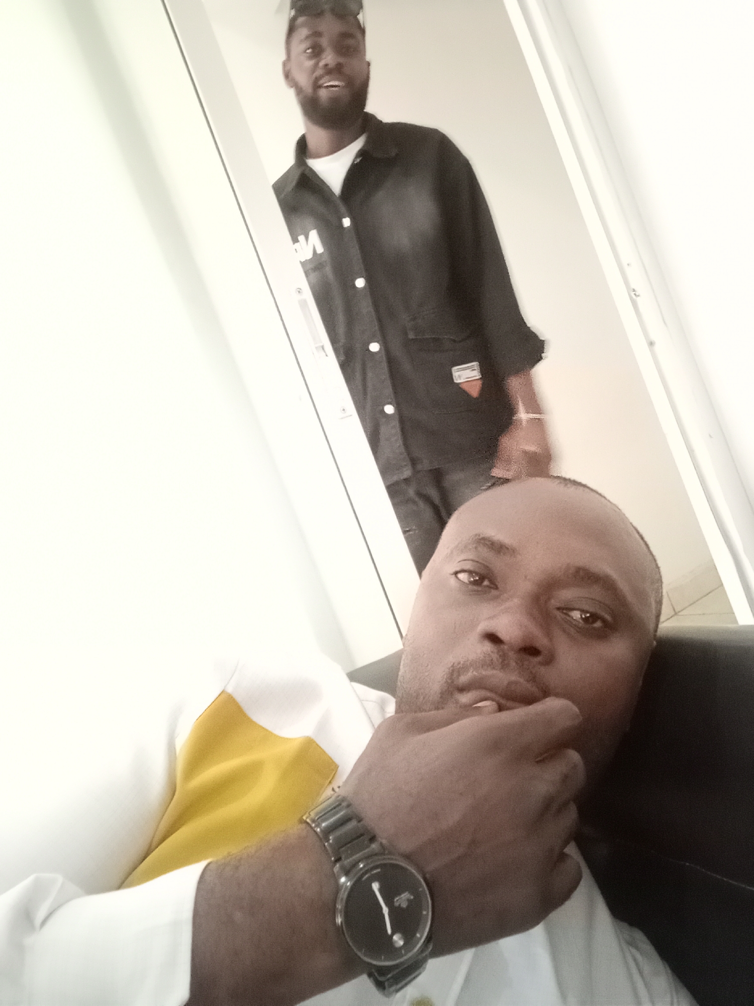 The image shows two Black African men indoors. The first man is closer to the camera, lying down or reclining in a dark-colored chair. He is wearing a white and yellow shirt and a wristwatch. His hand is near his mouth, and he is looking directly at the camera. The second man is standing in the background, appearing to be in a doorway. He is wearing a dark-colored shirt or jacket and dark trousers. The setting appears to be a room with light-colored walls and a white doorframe. The location is Gwarinpa, Nigeria.