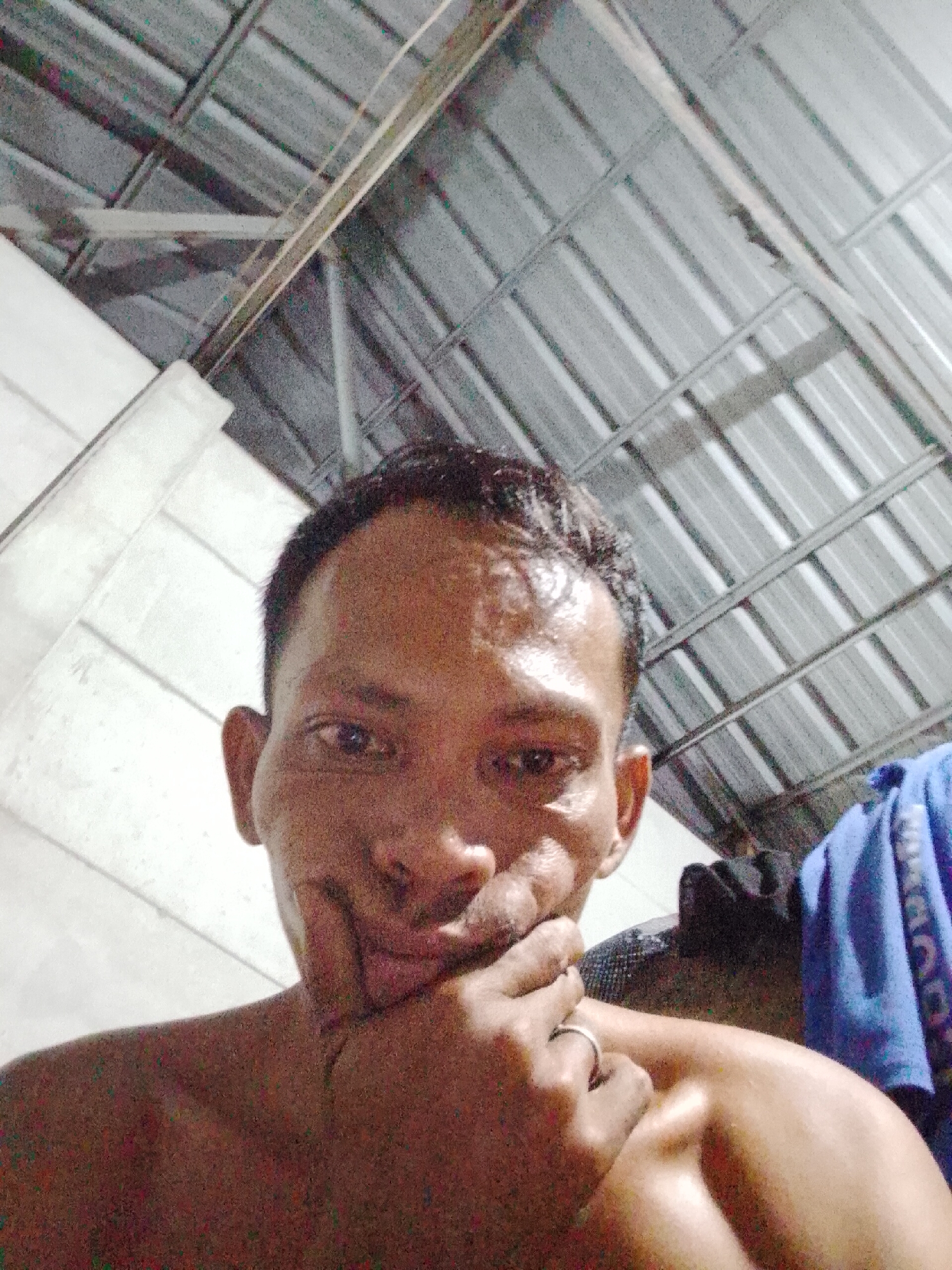 A shirtless man with short dark hair is posing for a selfie indoors. He is holding his chin with his hand, on which he is wearing a ring. The background includes white walls and a corrugated metal roof with visible support beams. A blue cloth is hanging in the background to the right. The image appears to be taken in Bukit Ketok, Indonesia.
