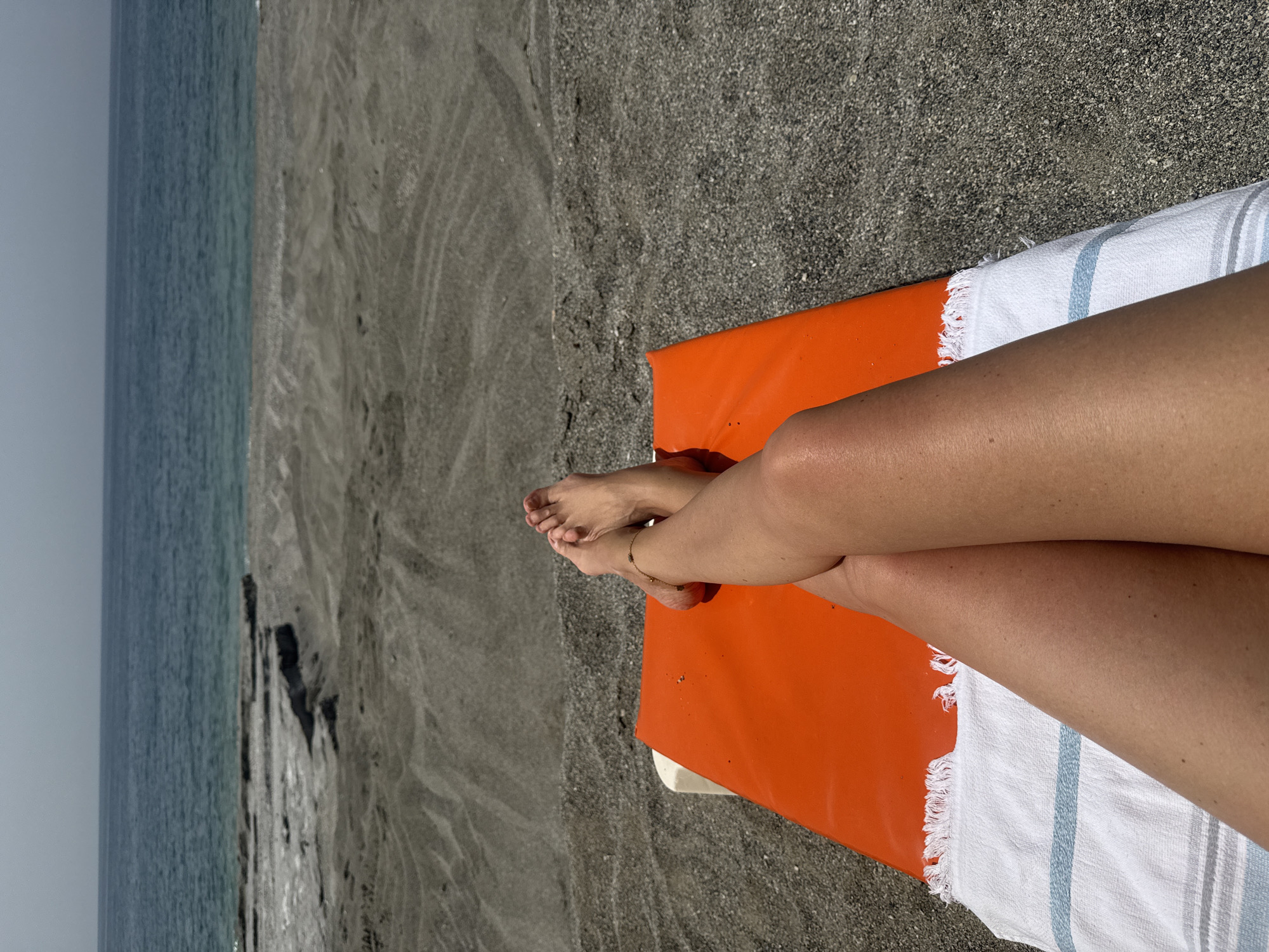 A person is lying on an orange lounge chair with a white and blue striped towel on a black sand beach. The person's legs are crossed, and they are wearing an anklet. The ocean is visible in the background, with some rocky formations near the shore. The sky is overcast. The location is Unknown, Unknown.