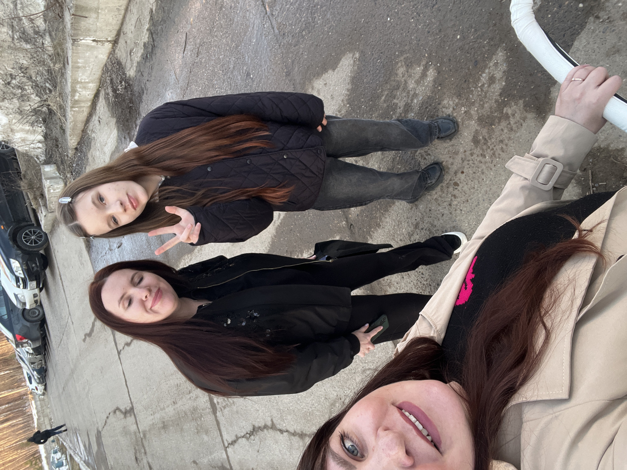 Three young women are posing for a selfie in Noviy Urengoy, Russia. The woman taking the selfie is wearing a beige coat. One of the women is wearing a black quilted jacket and jeans and holding up a peace sign. The other woman is wearing a black jacket and black pants, holding her phone in her hand. They are standing on a concrete surface. There are parked cars in the background.