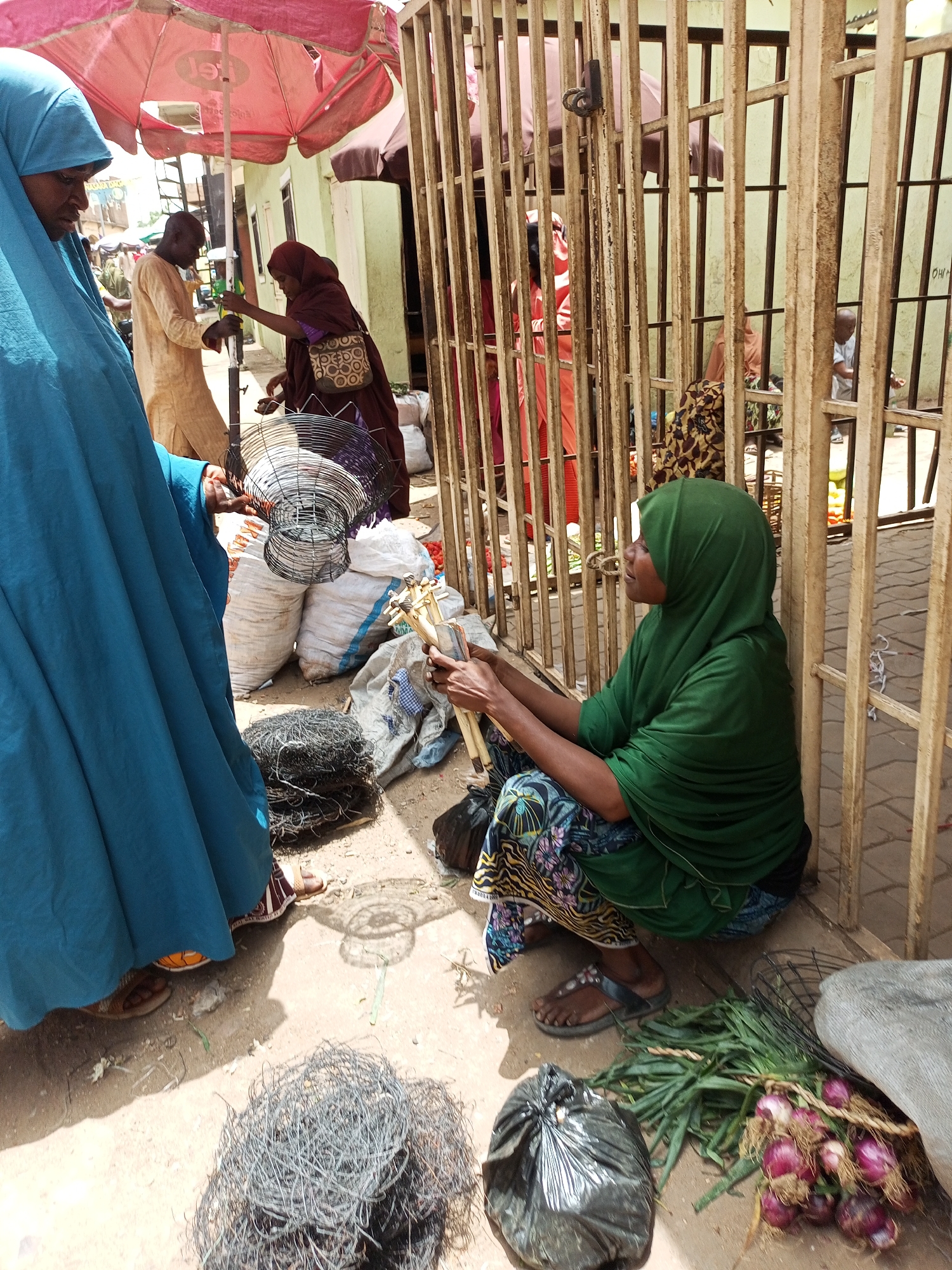 The image shows a street scene in Arawa, Nigeria. A woman in a green hijab and patterned skirt squats near a metal gate, holding wooden sticks. Nearby are onions and a black plastic bag. A woman in a blue burqa stands facing her, holding a wire basket. Other people and shops are visible in the background, beneath red umbrellas. The environment appears to be a marketplace, with goods displayed on the ground.